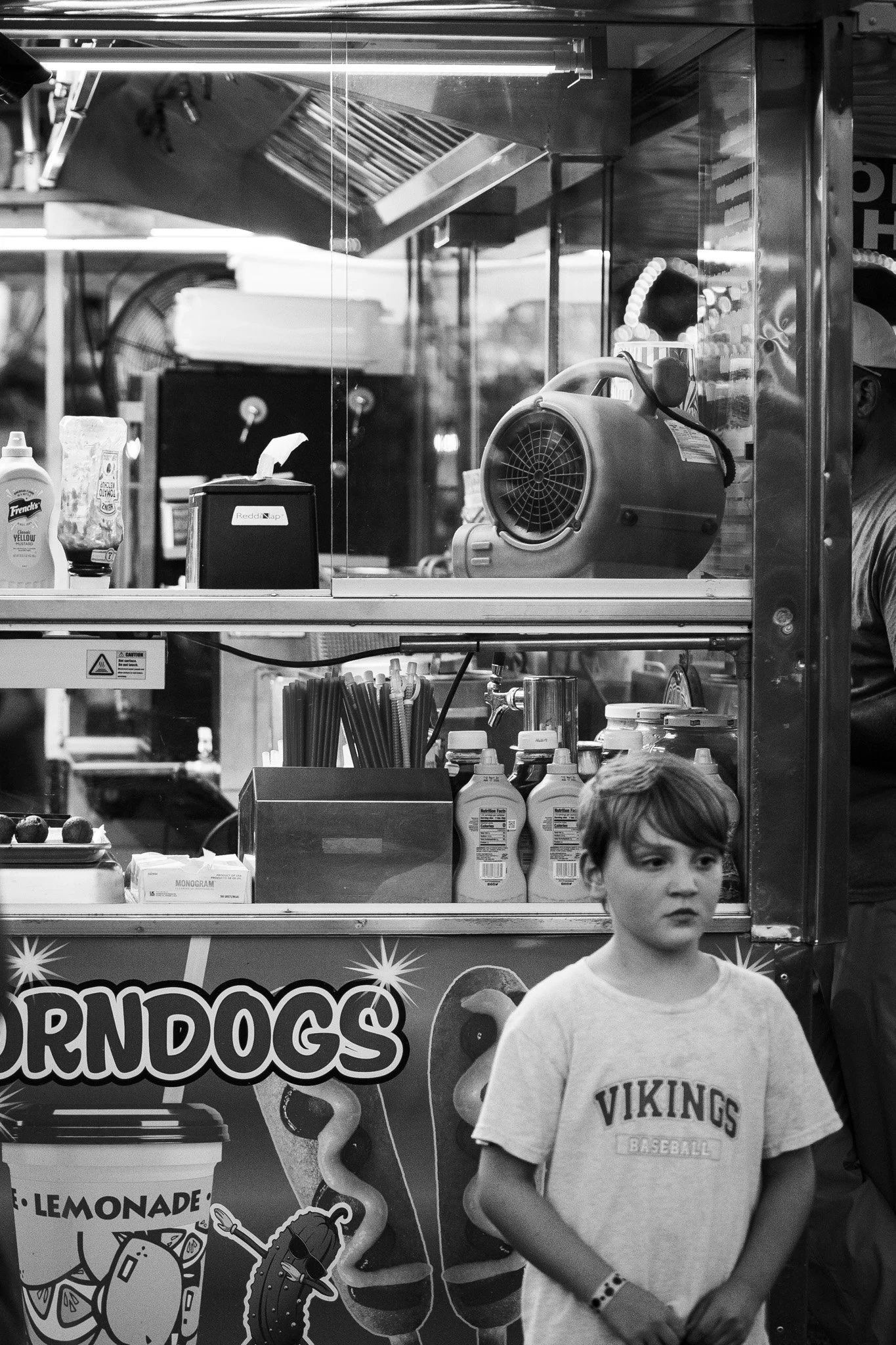 A young boy wearing a t-shirt with 'VIKINGS BASEBALL' printed on it standing in front of a food stand with a sign that says 'CORN DOGS' visible. The stand has various items including bottles, a fan, and a large fan heater or blower.