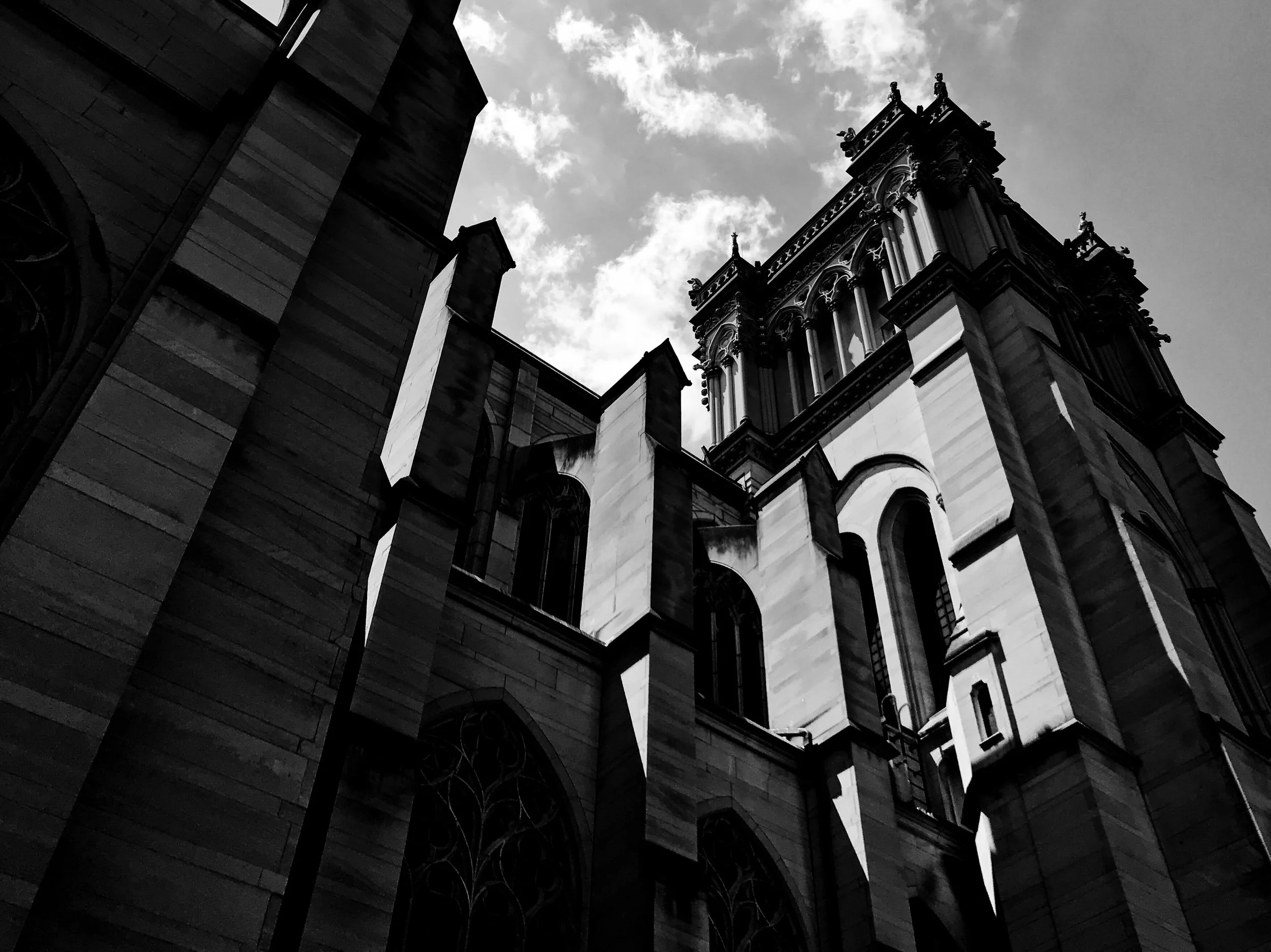 A black and white photo of a historic or Gothic-style building with intricate architectural details, pointed arches, and tall spires, taken from a low angle against a partly cloudy sky.