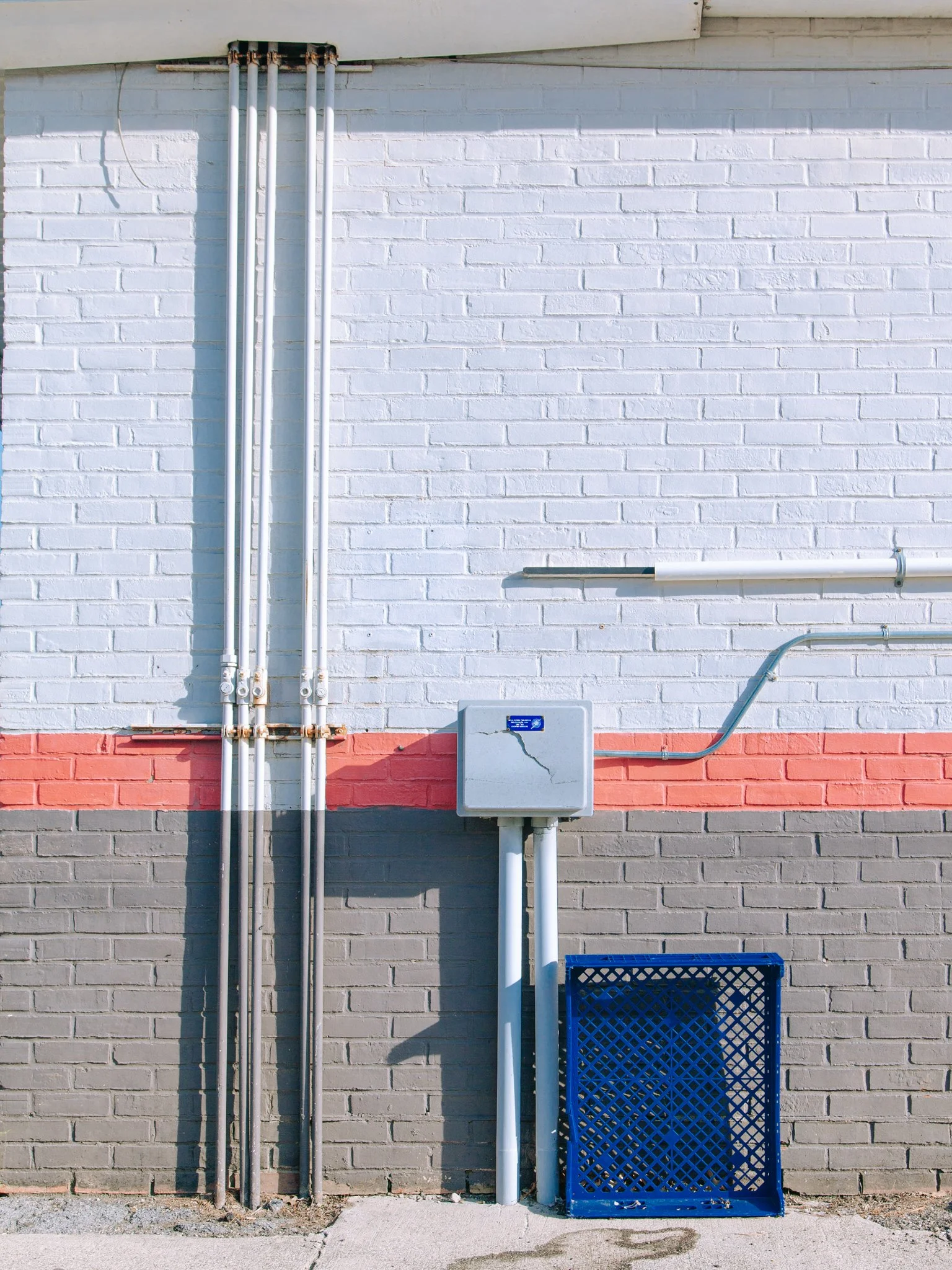A brick wall painted white with a red stripe near the bottom. There are four vertical pipes on the left, a small utility box in the center, and a blue plastic crate on the right.