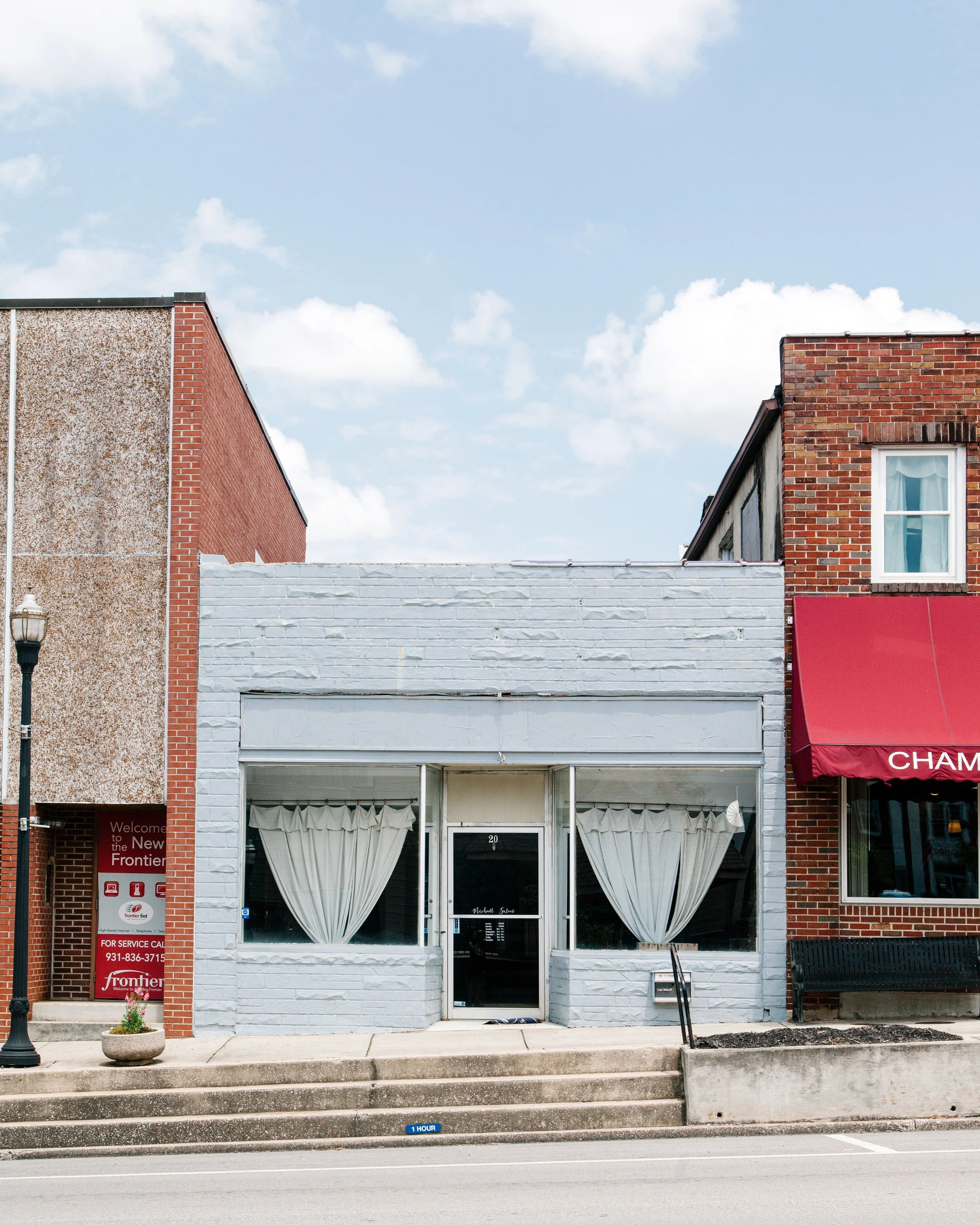 Facade of a small storefront with white-painted brick wall, large front window with curtains, black door, and a red awning reading "CHAM". Sidewalk steps lead up to the entrance with an 