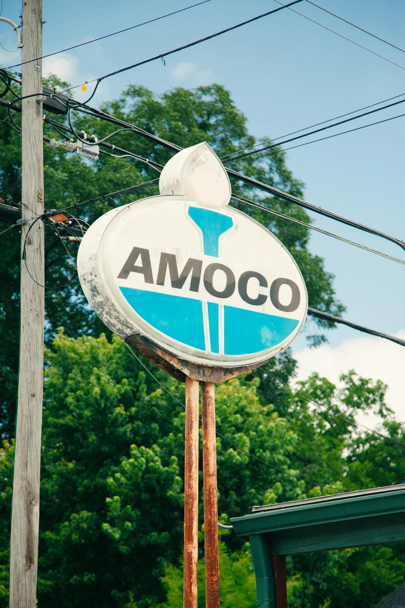 A vintage AMOCO gas station sign with a partly cloudy sky, a utility pole with wires, green trees in the background, and part of a building's roof in the foreground.