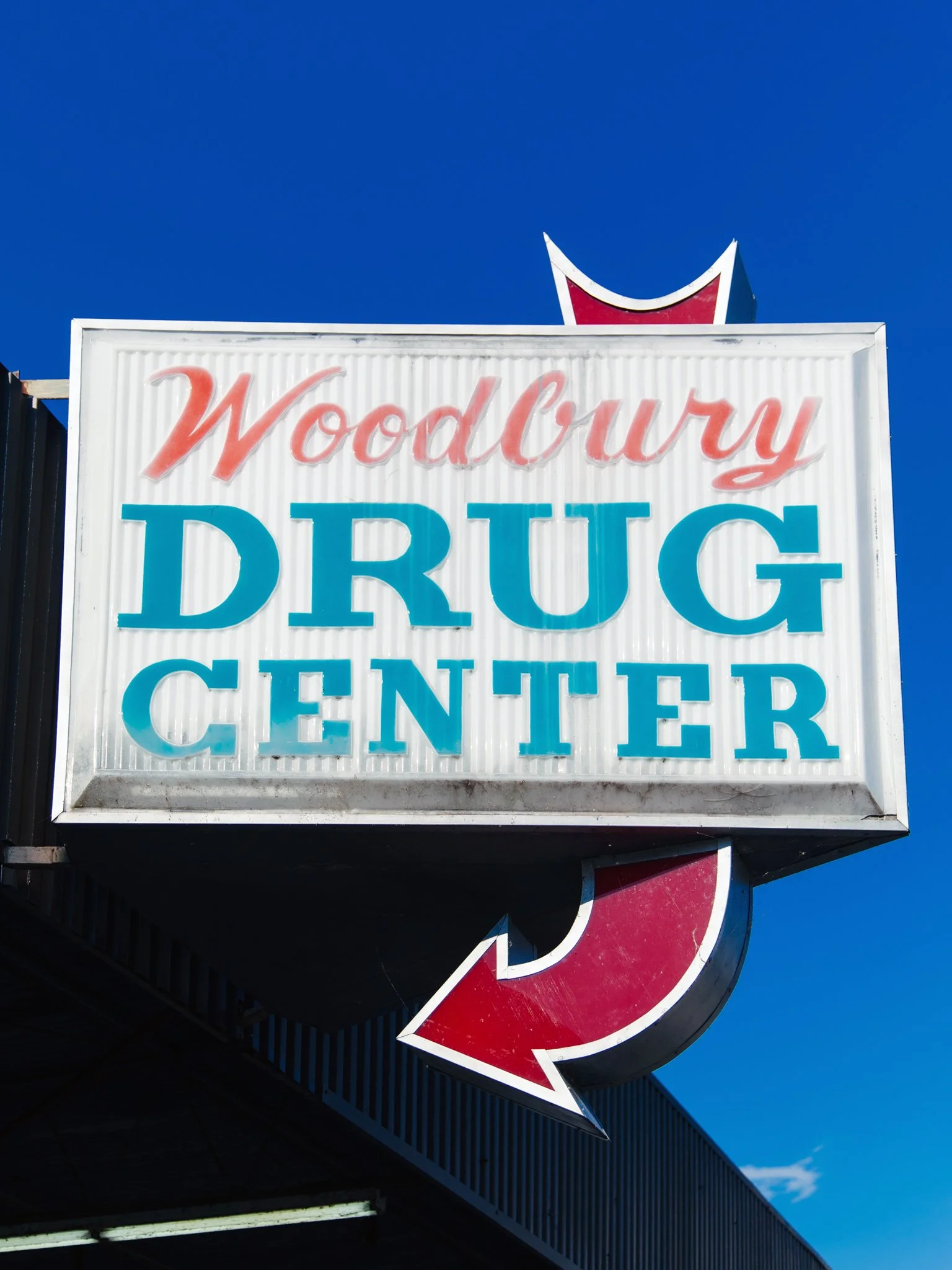 Sign for Woodbury Drug Center with a red and white arrow pointing to the left, mounted on a building against a bright blue sky.