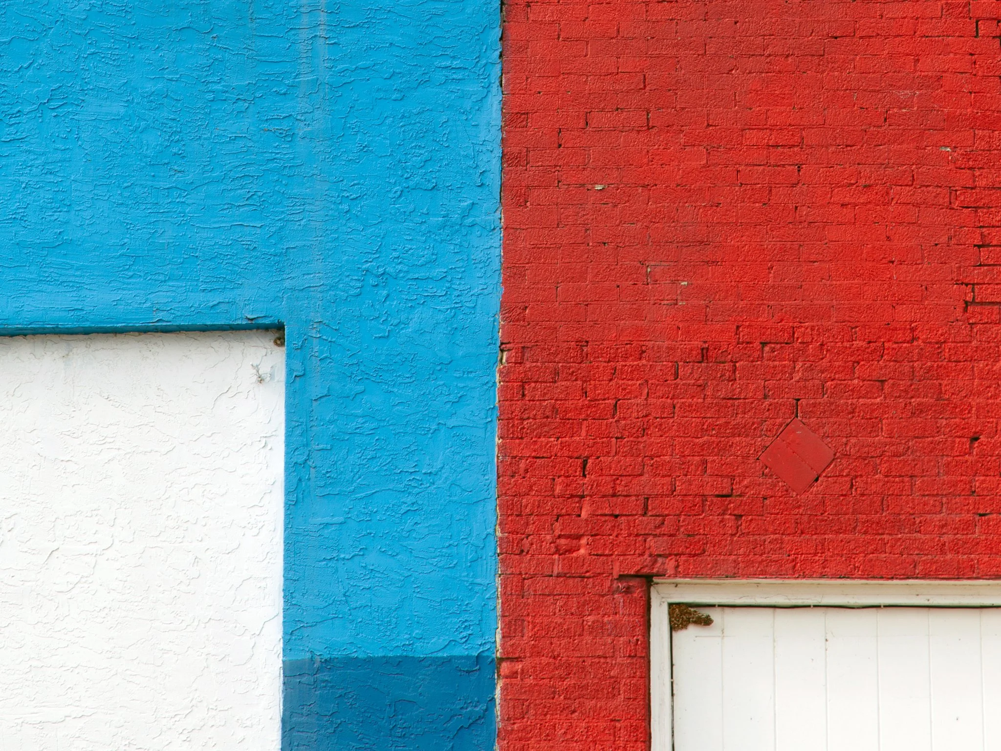 Close-up of a wall divided into three sections: the left section is white with a textured surface, the middle section is blue with a rough, textured paint, and the right section is red brick with a small window opening at the bottom.