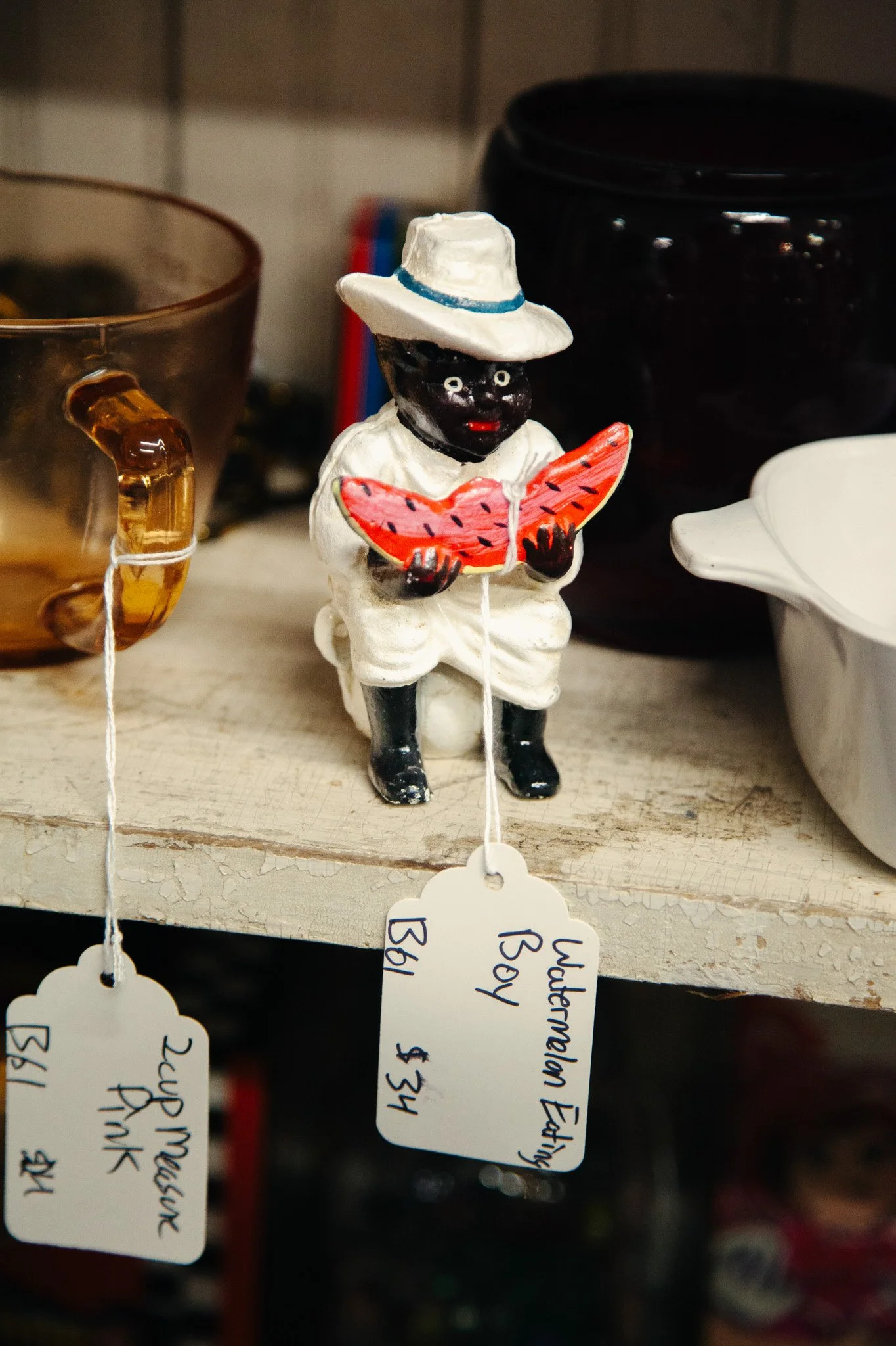 A decorative figurine of a person with dark skin and white clothes, wearing a white hat with a blue band, holding a pink and orange watermelon slice, on a shelf with other items in the background.
