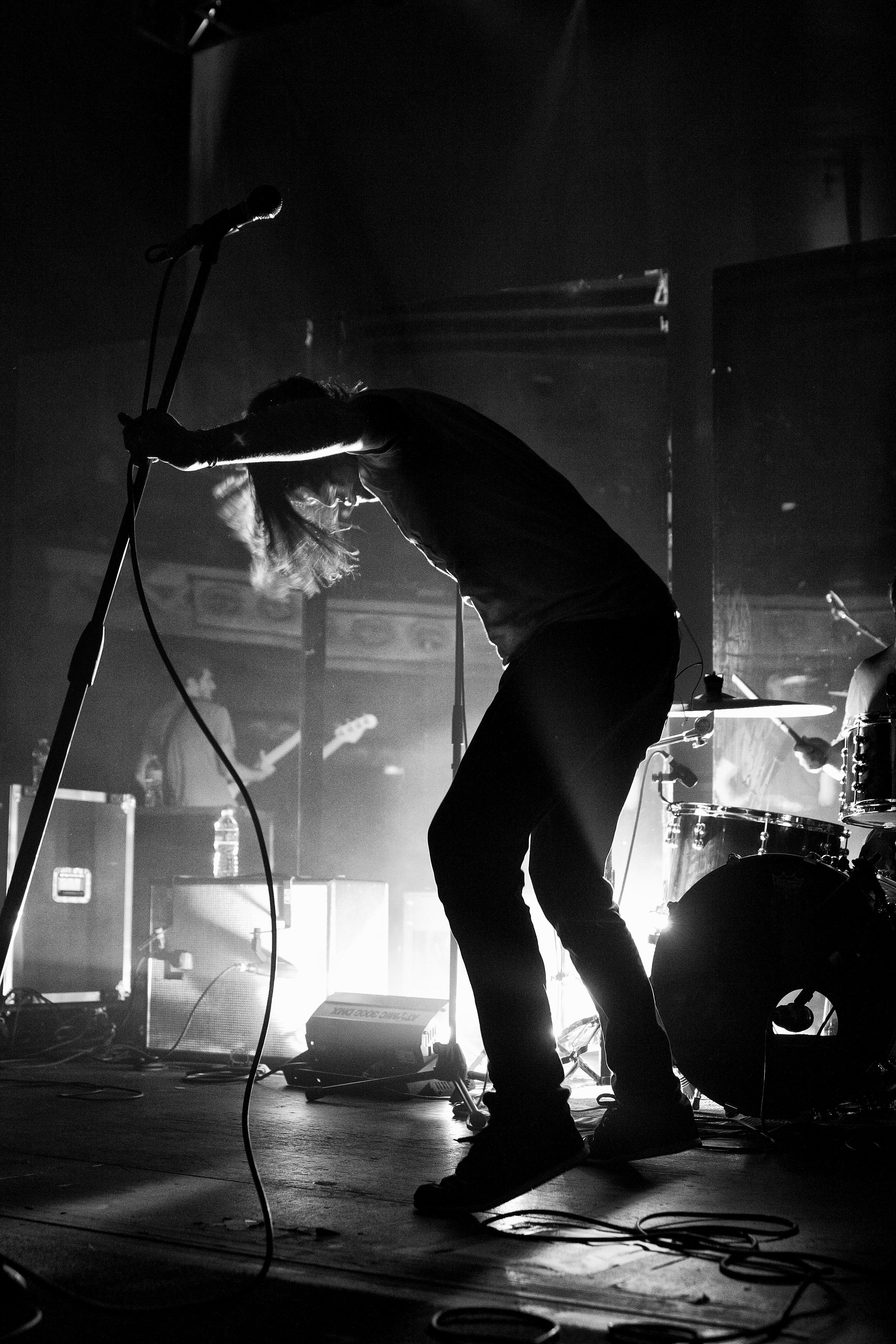 A musician performing on stage with a microphone, in a dimly lit, black-and-white setting, surrounded by musical equipment.