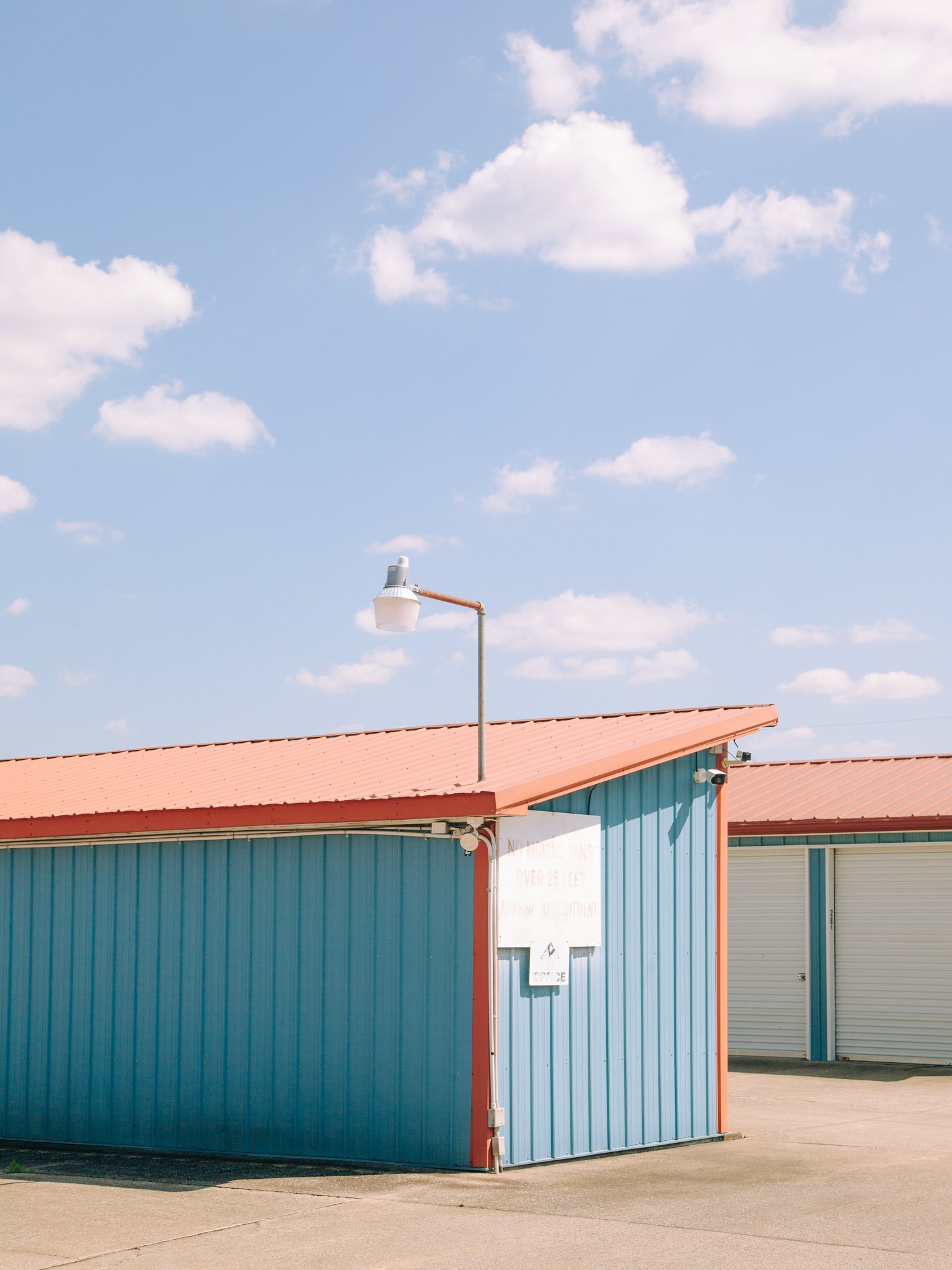A blue building with a red roof and a white sign, next to a garage door. An outdoor light fixture is mounted on a pole in front. The sky is partly cloudy with white clouds.