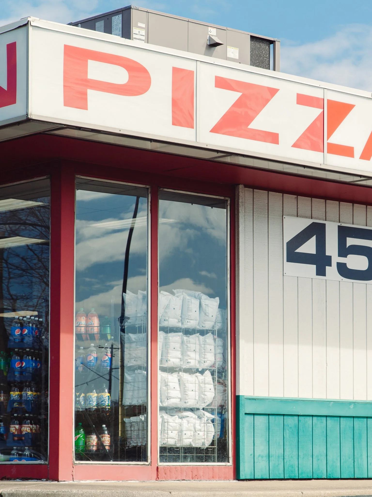 Exterior of a convenience store with a sign that reads 'PIZZA'. The store has large glass windows displaying various beverages, including Pepsi, 7UP, Mountain Dew, and other soft drinks. The building has a white and turquoise color scheme with red ac