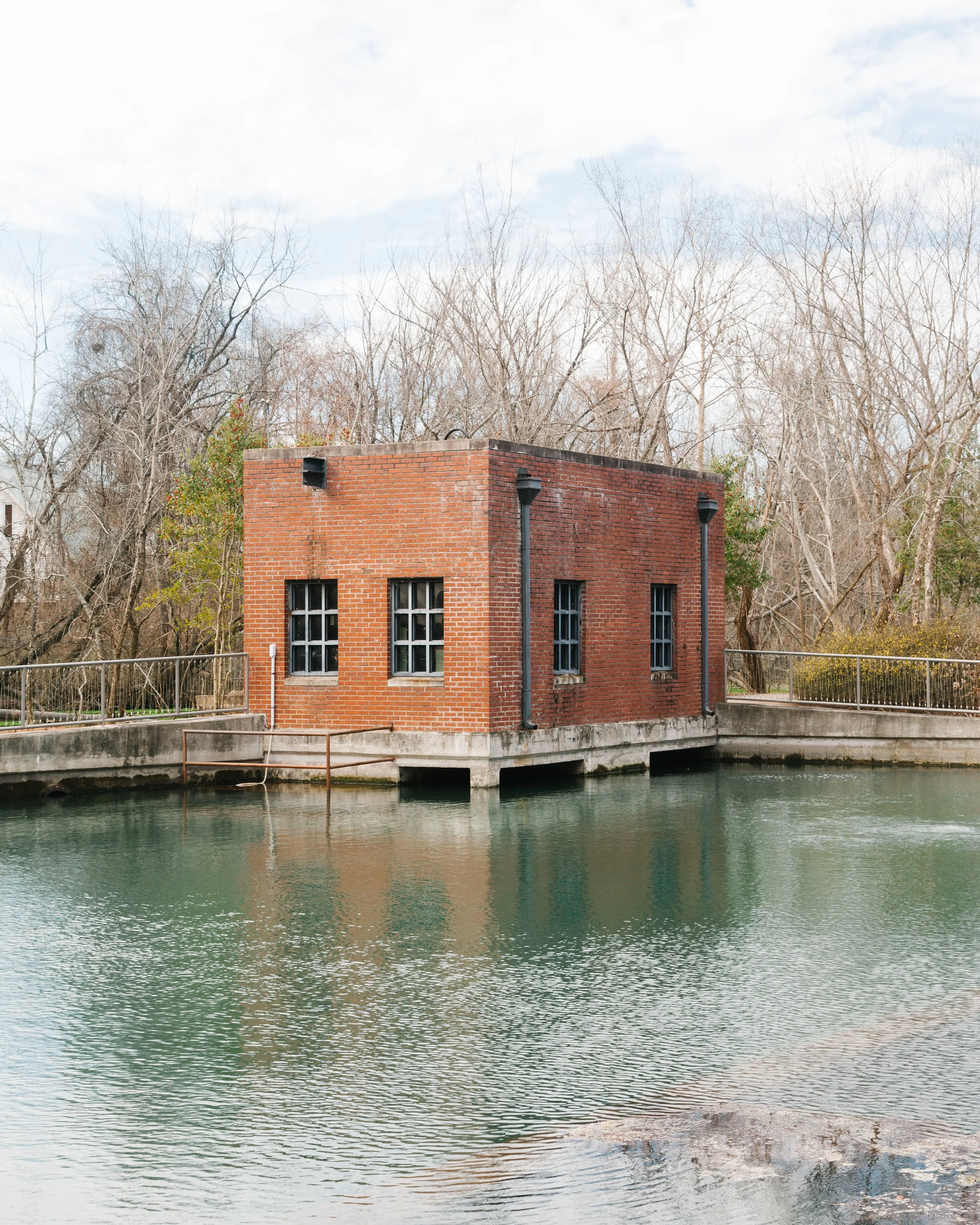 A brick building on a small bridge over a calm body of water with leafless trees in the background