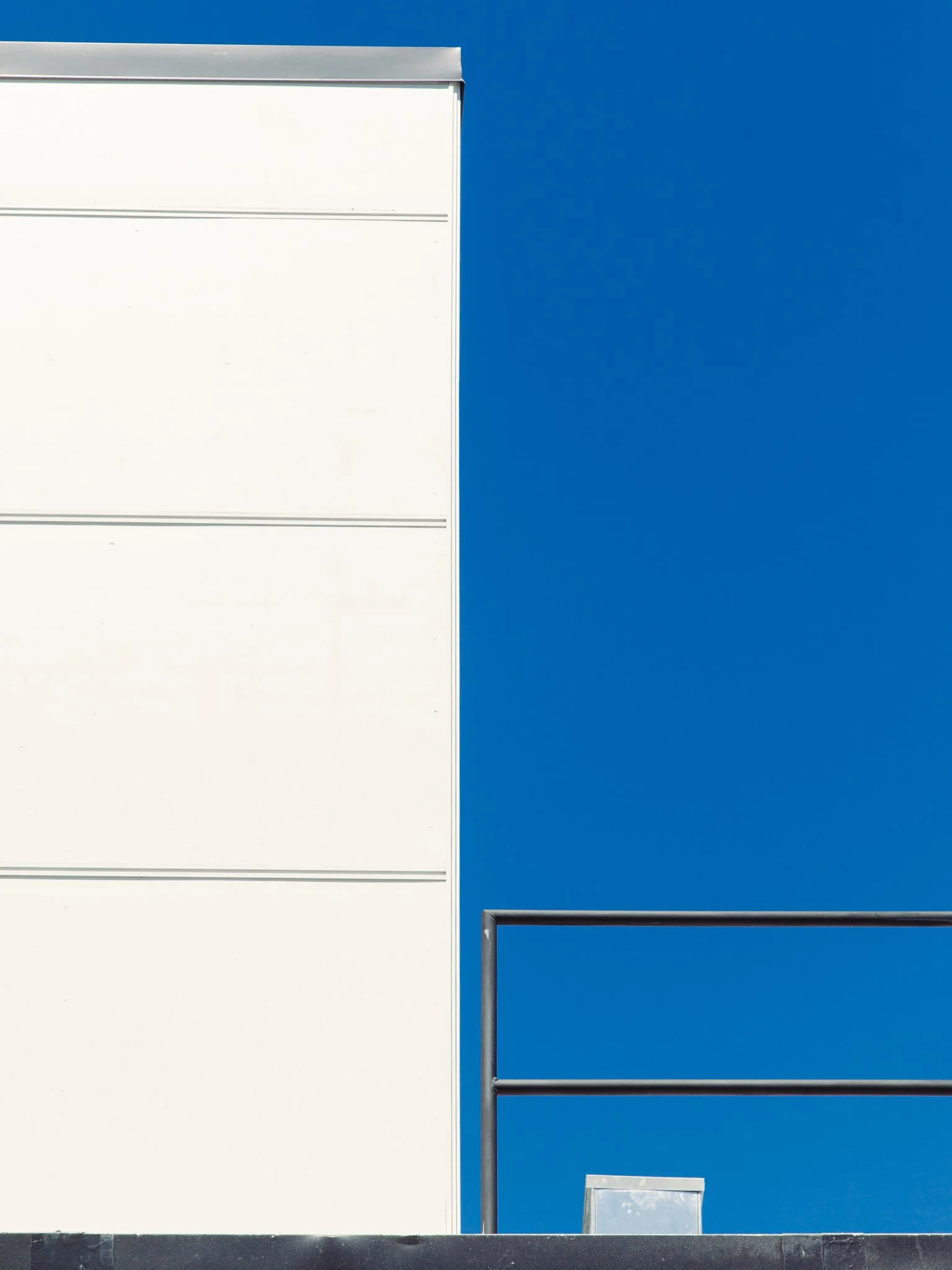 Close-up of the corner of a modern building with white and blue panels, metal framing, and a vent at the bottom, against a bright blue sky.