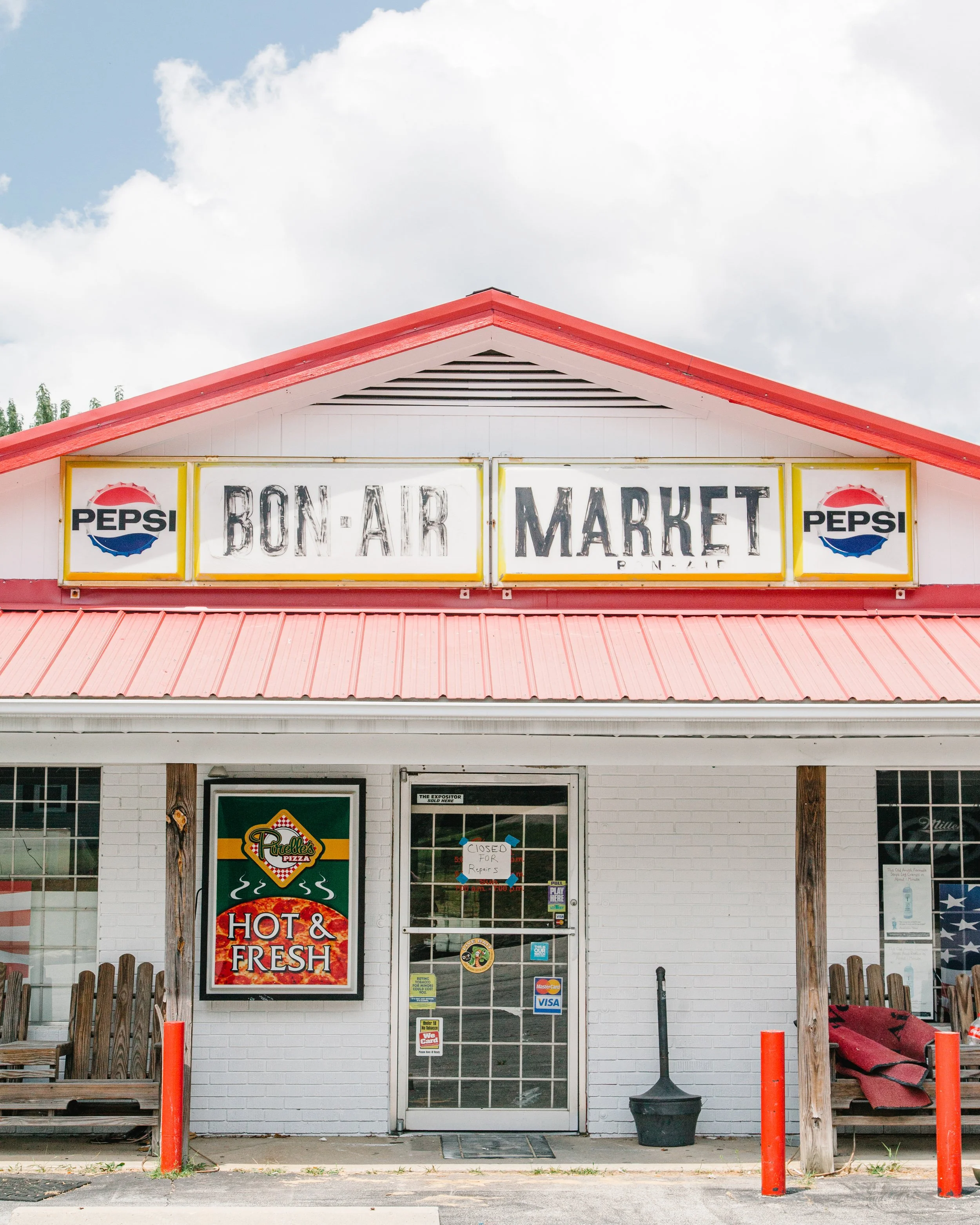 Front view of a small building with a red roof, white brick walls, and a door with stickers. A sign above the door reads 'Bon-Air Market,' and there is a Pepsi advertisement on the left side of the sign. A bench is on the left, and a wheelchair ramp 