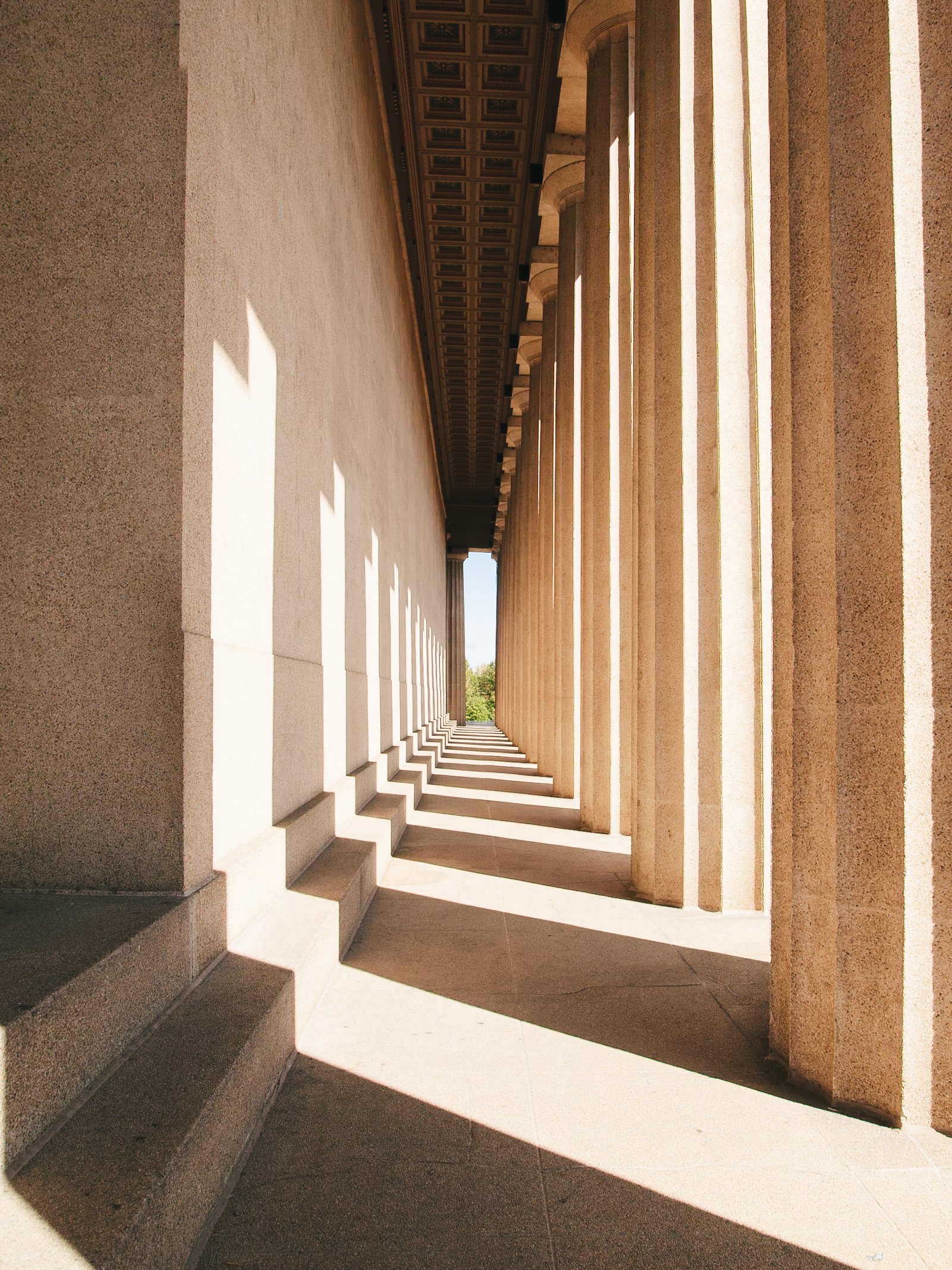 Sunlit neoclassical architectural colonnade with stairs, columns, and decorative ceiling panels.