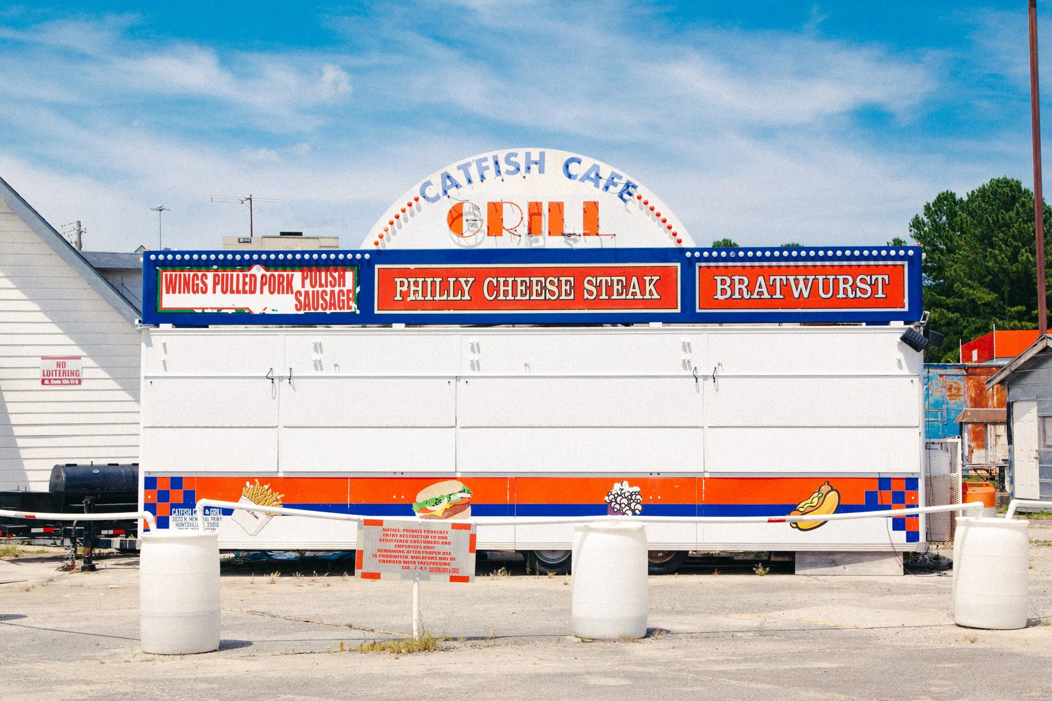 Front view of a vintage-style fast food stand with signage for Catfish Cafe Grill, offering chicken wings, pulled pork, polish sausage, Philly cheesesteak, and bratwurst, with colorful illustrations of food items on the lower panel.