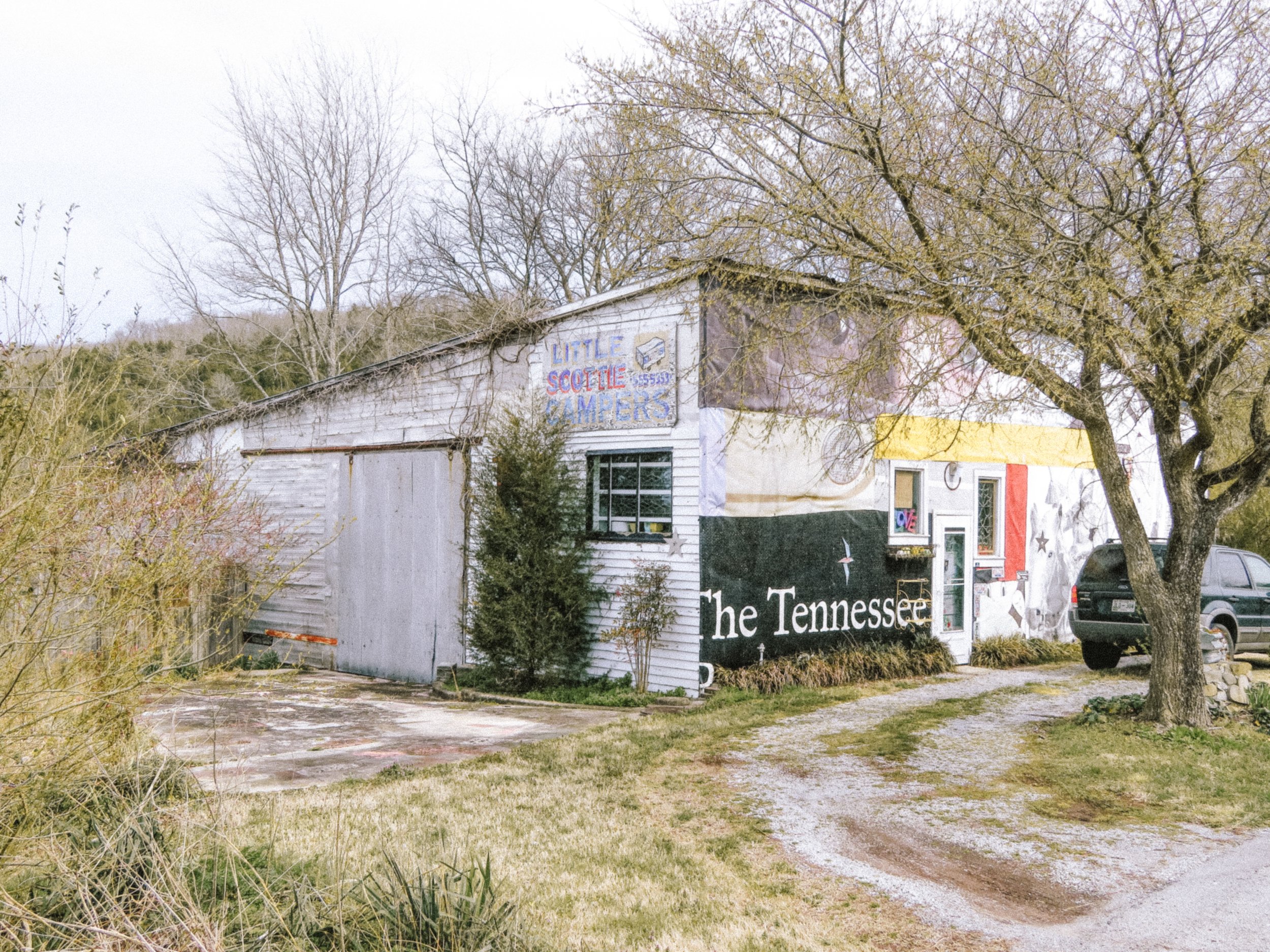 An old, weathered white building with a sign that reads "LITTLE SCOTTY CAMPERS" is partially obscured by leafless trees. The building has a mural with the words "the Tennessee" visible at the bottom. The scene is set in a rural area with a dirt path 