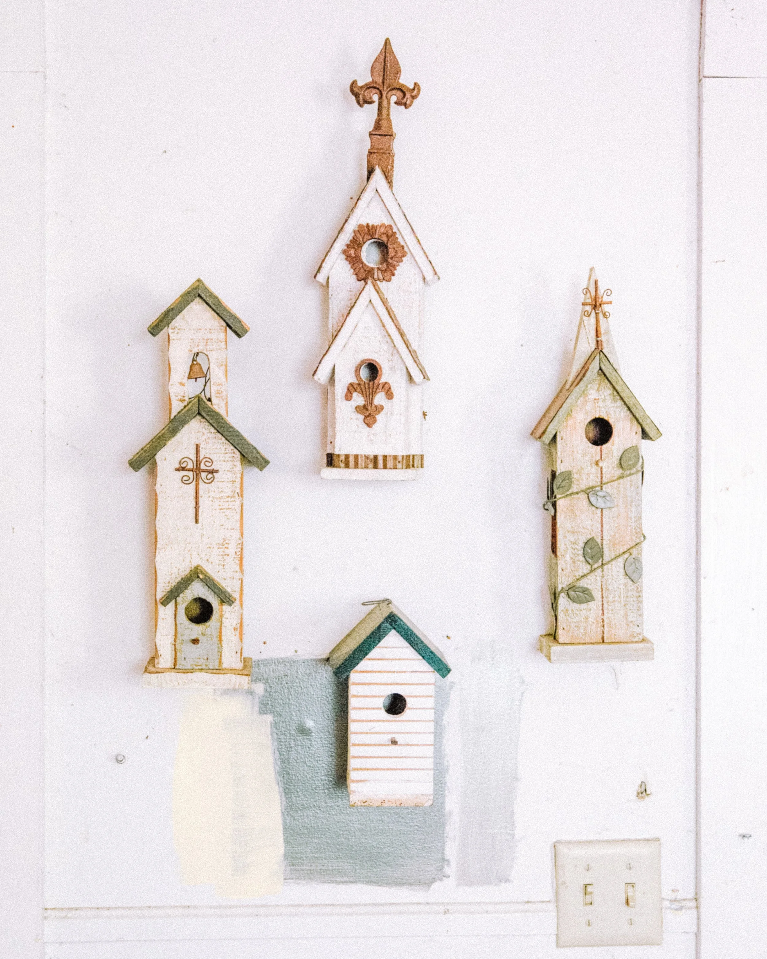 Three decorative miniature birdhouses on a white wall with an electrical outlet below.