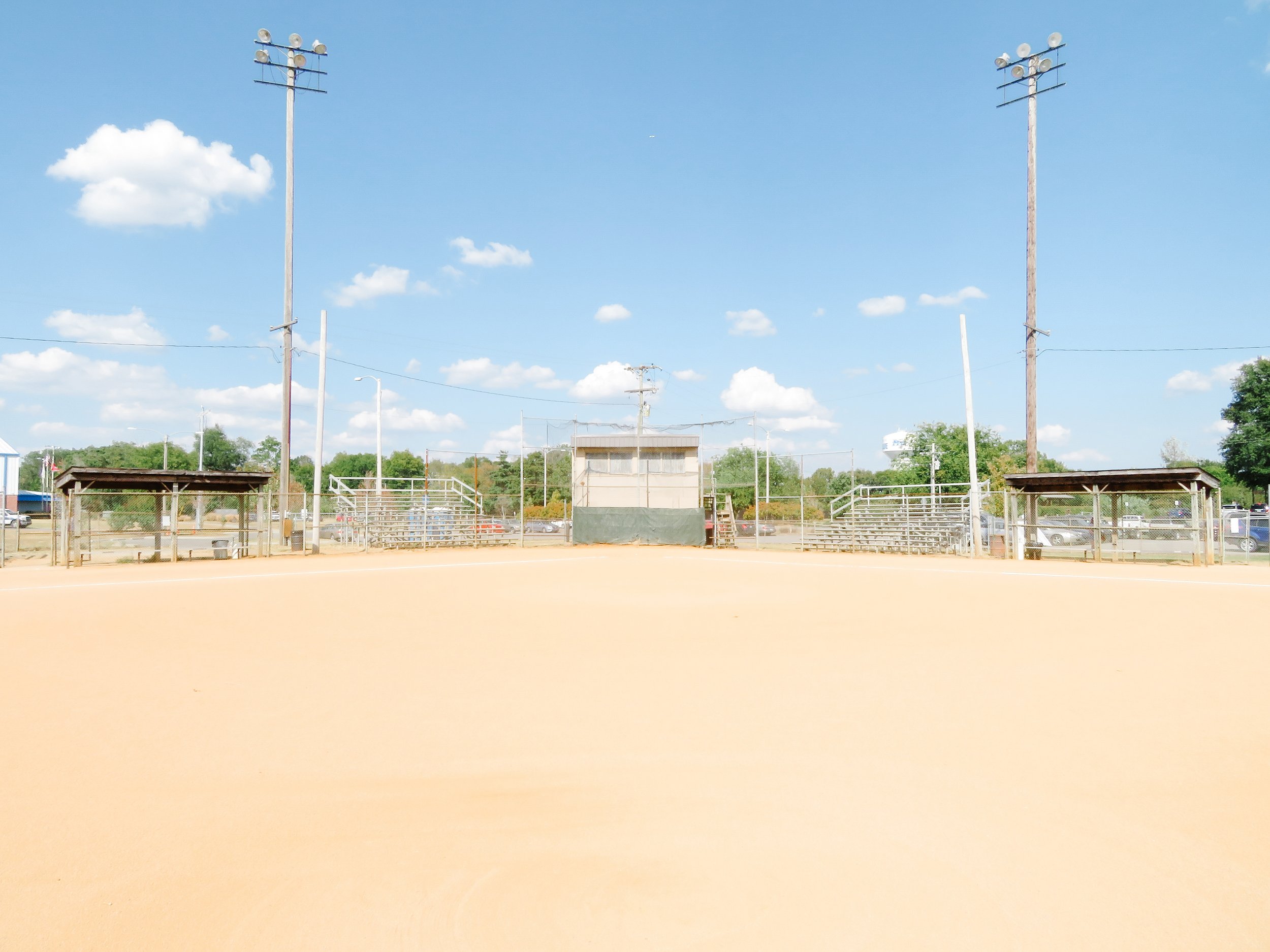 Baseball field with bleachers, a small building, streetlights, and trees in the background under a blue sky with scattered clouds.