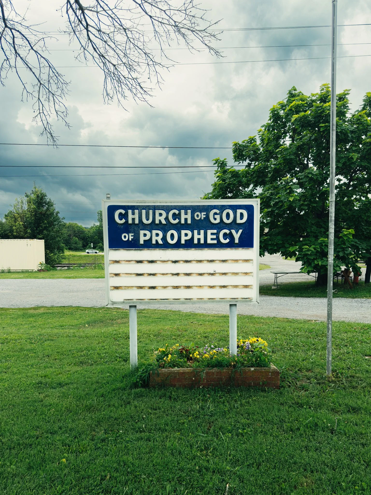 A sign reading 'Church of God of Prophecy' in a grassy area with trees and cloudy sky in the background.