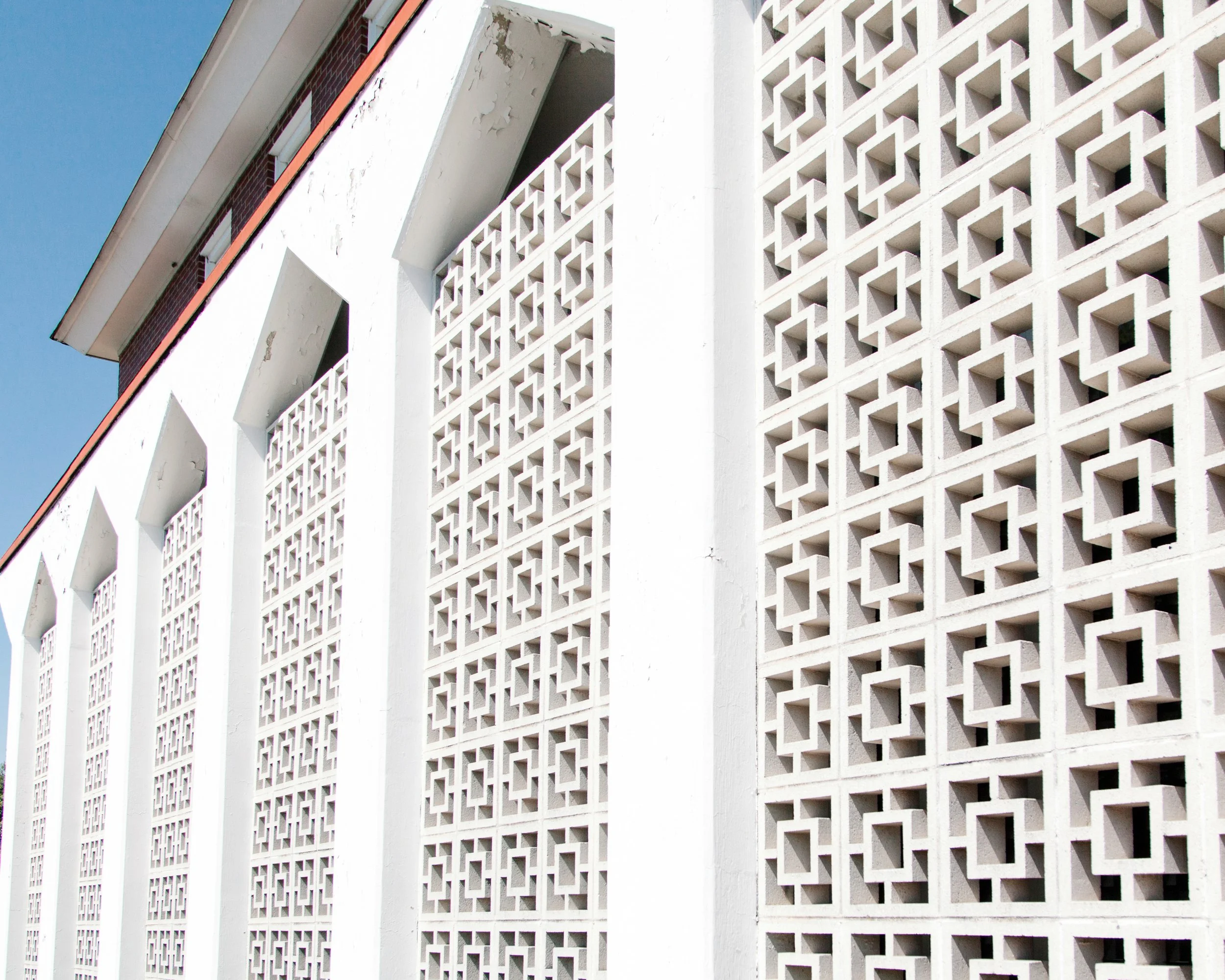 A close-up view of a white concrete block wall with geometric cut-out patterns, part of a building exterior against a blue sky, with a section of a terracotta-tiled roof.