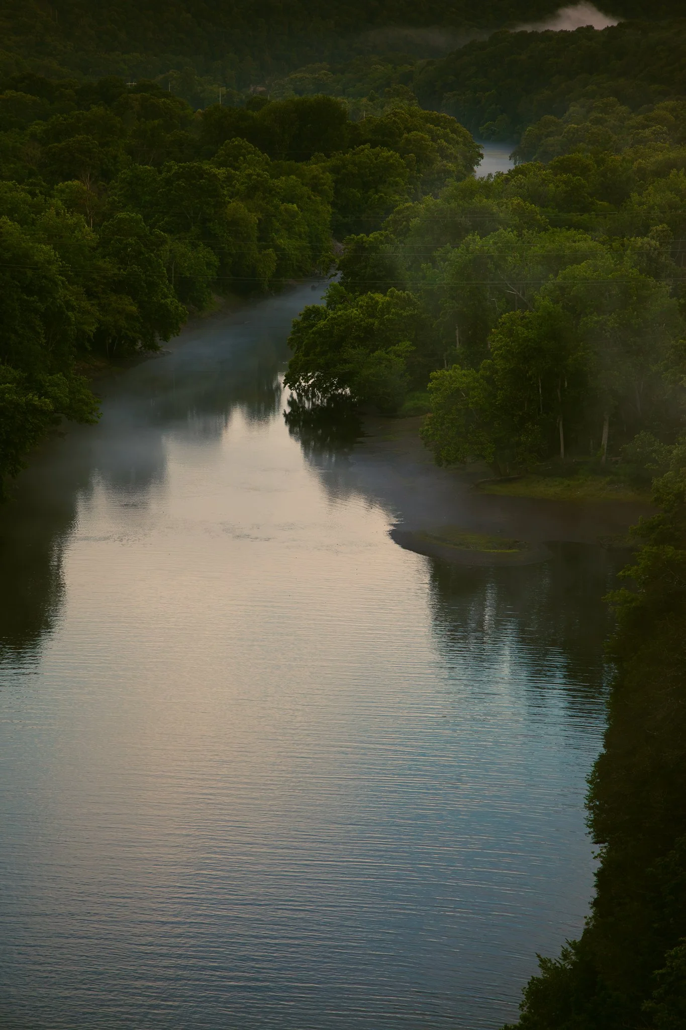 A river flowing through a dense, green forest with hills in the background, under a partly cloudy sky.