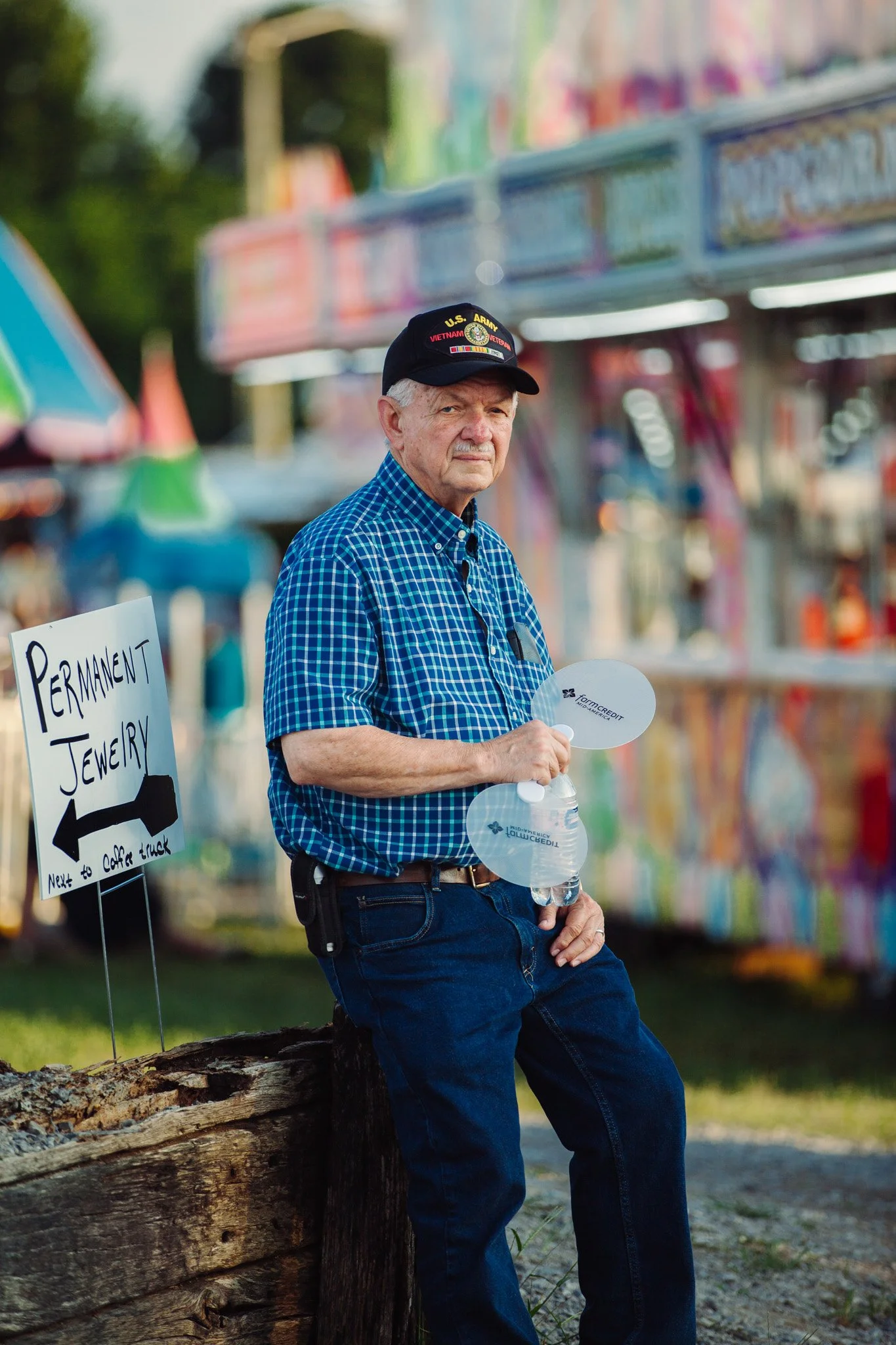 An elderly man wearing a U.S. Army Vietnam veteran cap is standing outdoors in front of a amusement park or carnival. He is holding a water bottle and leaning on a wooden log, with game booths or rides blurred in the background.