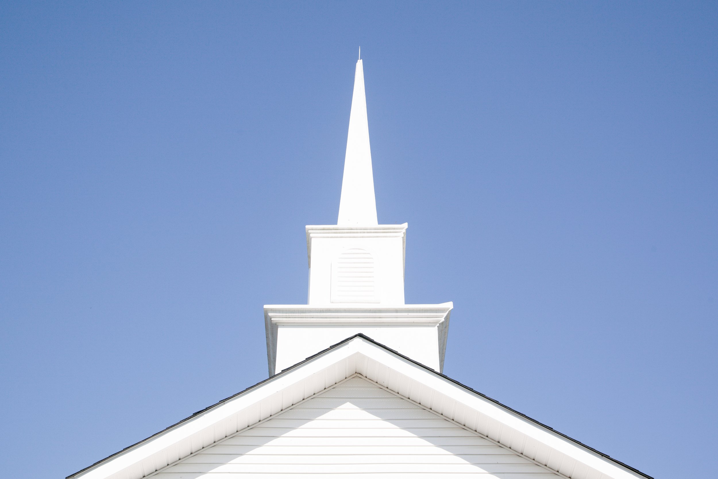 A white church steeple against a clear blue sky, with architectural details of the church's roof at the bottom.