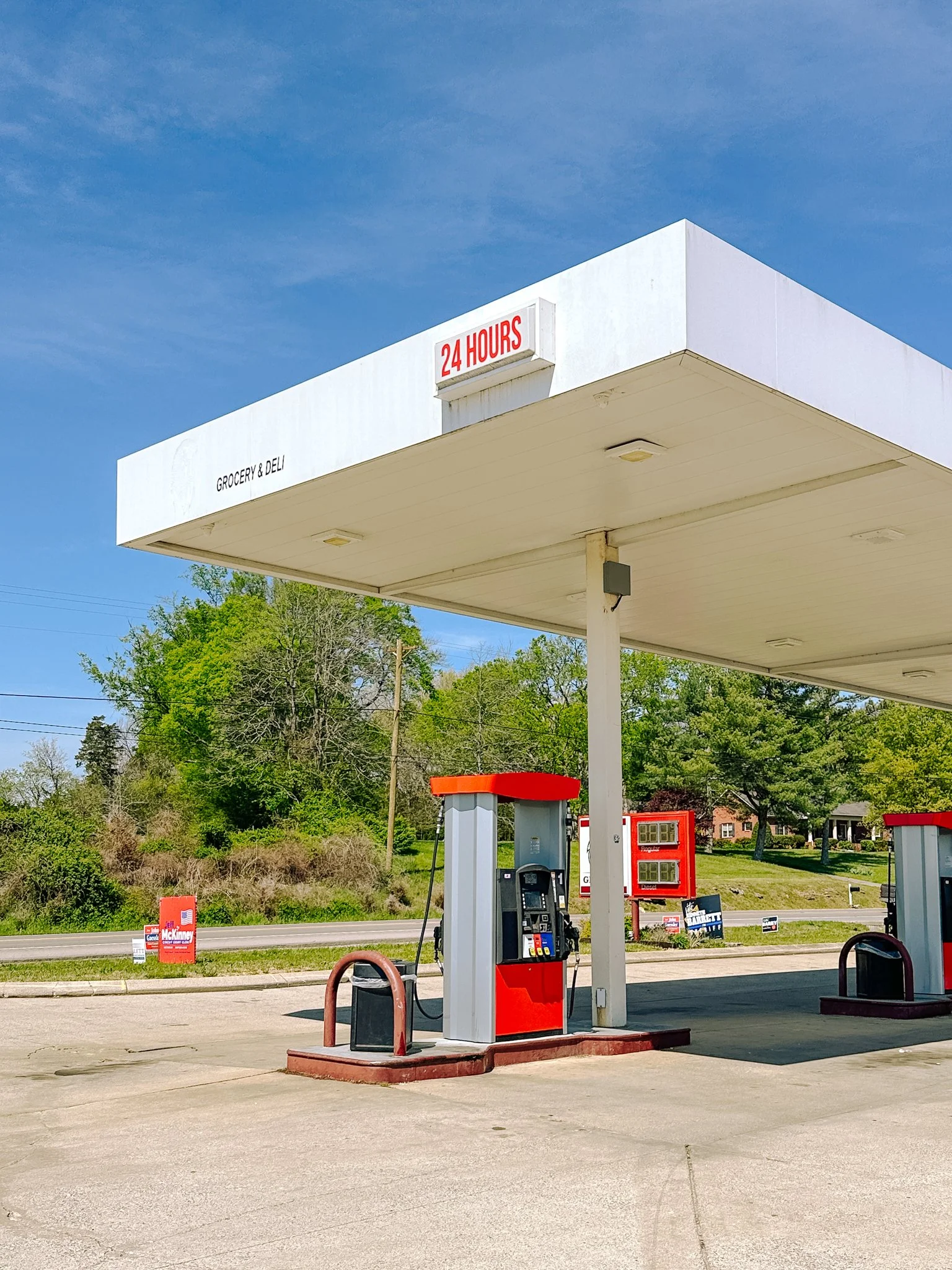 A gas station with a fuel pump, a red and gray color scheme, and a white canopy with a sign that says '24 HOURS.' In the background, there are trees, a grassy area, and houses.