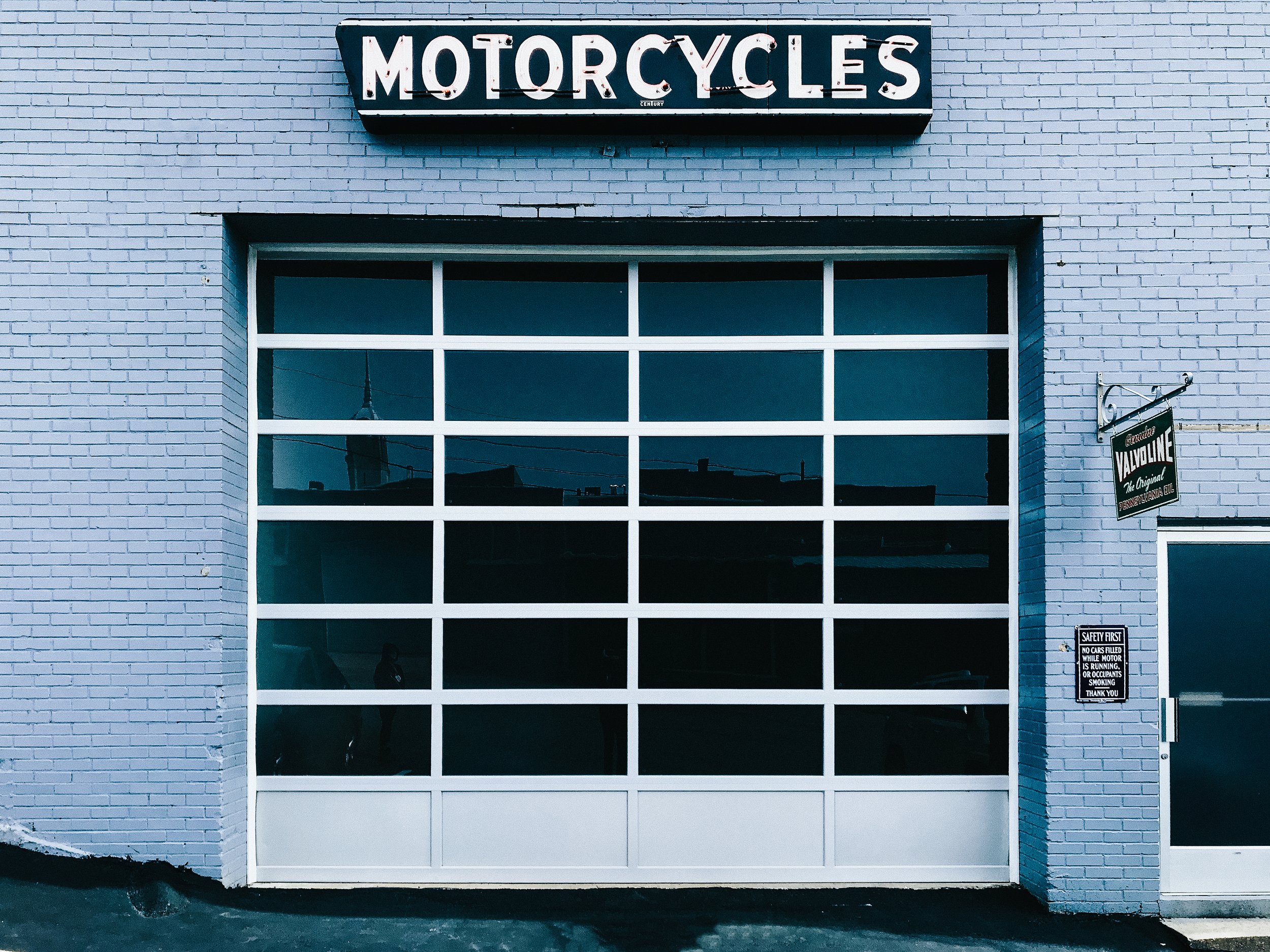 A large white brick building with a garage door window and a "Motorcycles" sign above it. There are safety notices on the wall next to the door.