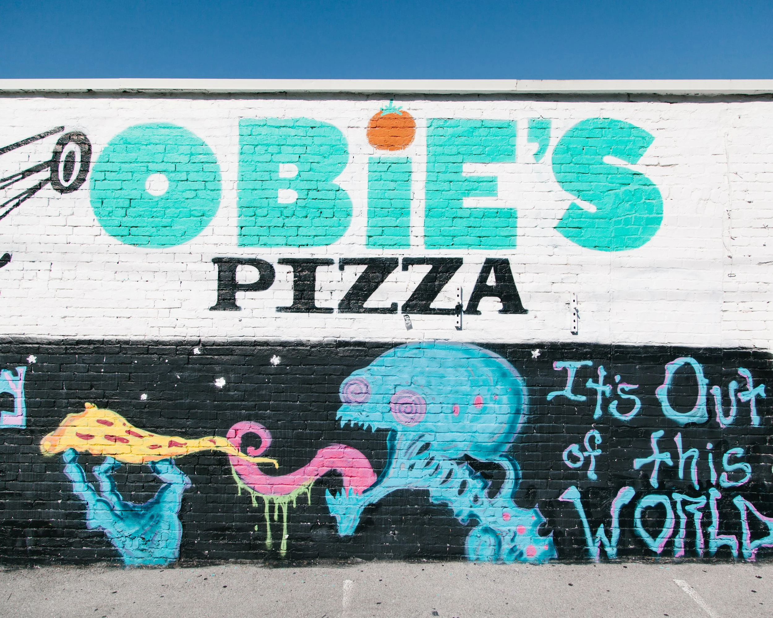 Colorful street art on a brick wall featuring the words 'O'Brien Pizza' with a little orange tomato above the '+', and a blue creature with pink heart-shaped eyes holding a yellow slice of pizza.