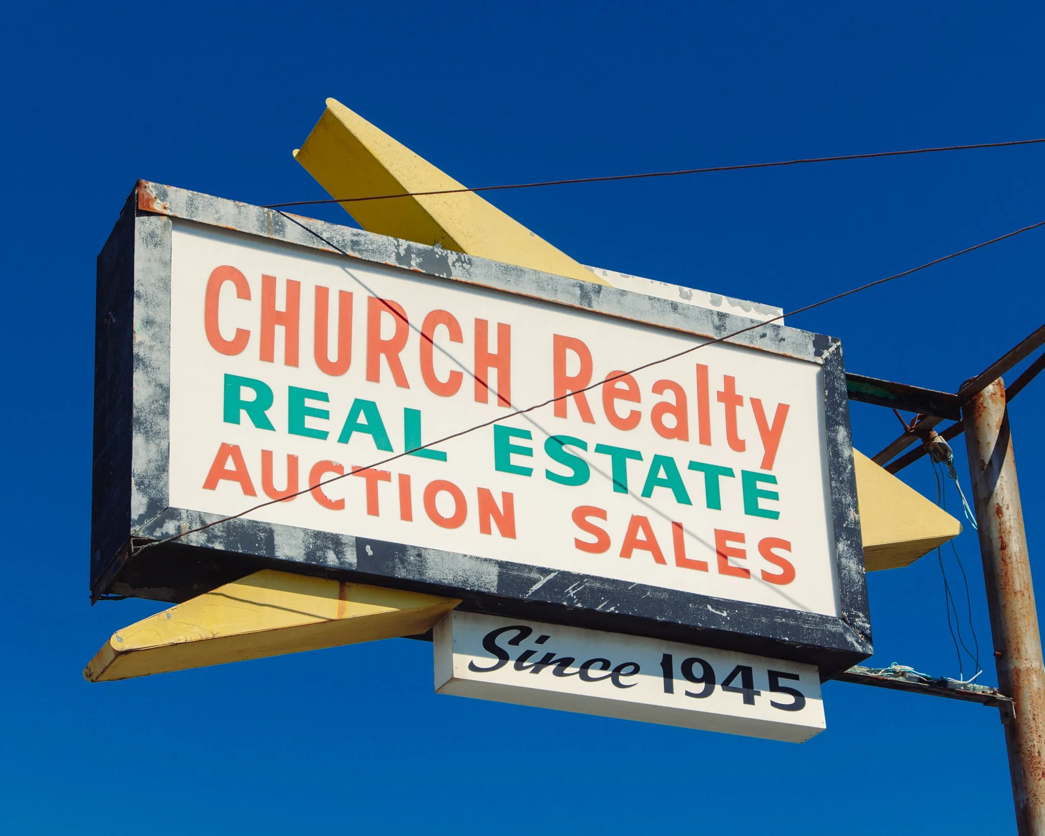 An outdoor street sign with multiple layers, stating 'Church, Realty, Real Estate, Auction Sale,' and indicating the street has been there since 1945, under a clear blue sky.