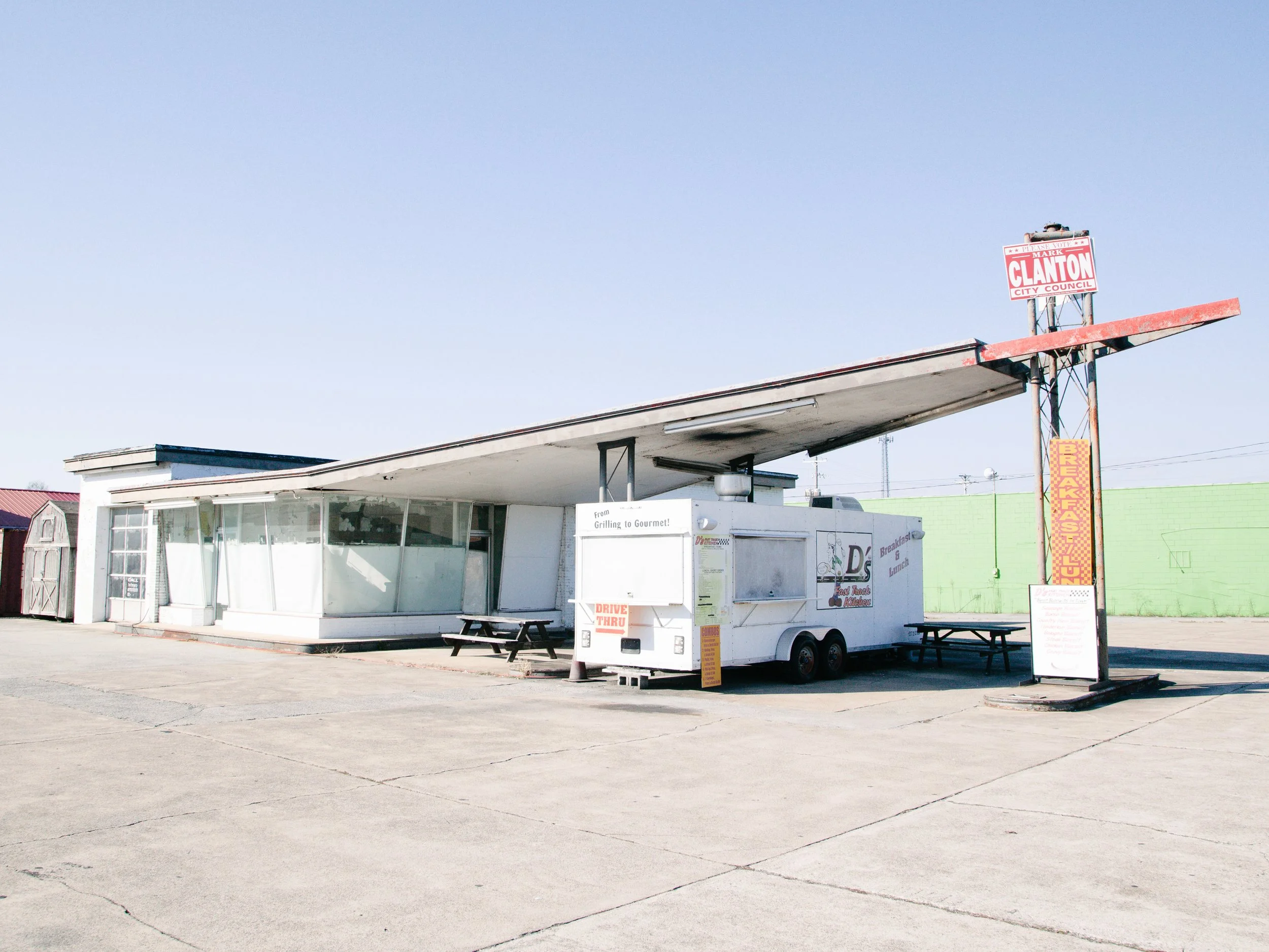 A food truck with a white exterior and large windows parked in an empty lot, with a sign advertising breakfast and lunch options, and a small picnic table nearby, set against a clear blue sky.