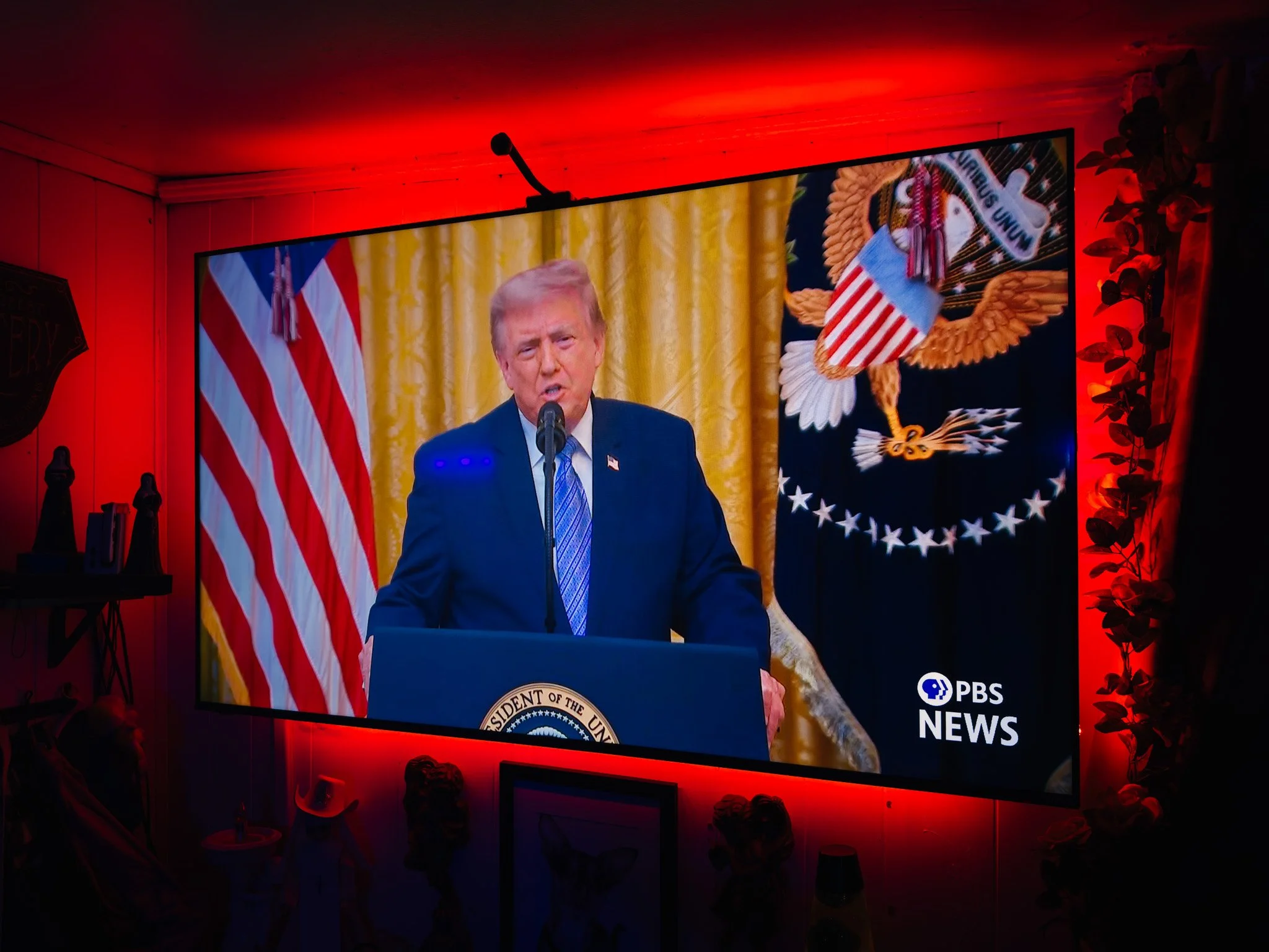 Image of former President Donald Trump speaking at a podium with a presidential seal in front of a yellow curtain. Behind him are American flags and a large eagle presidential seal. The scene is illuminated with red lighting, and there are people in 