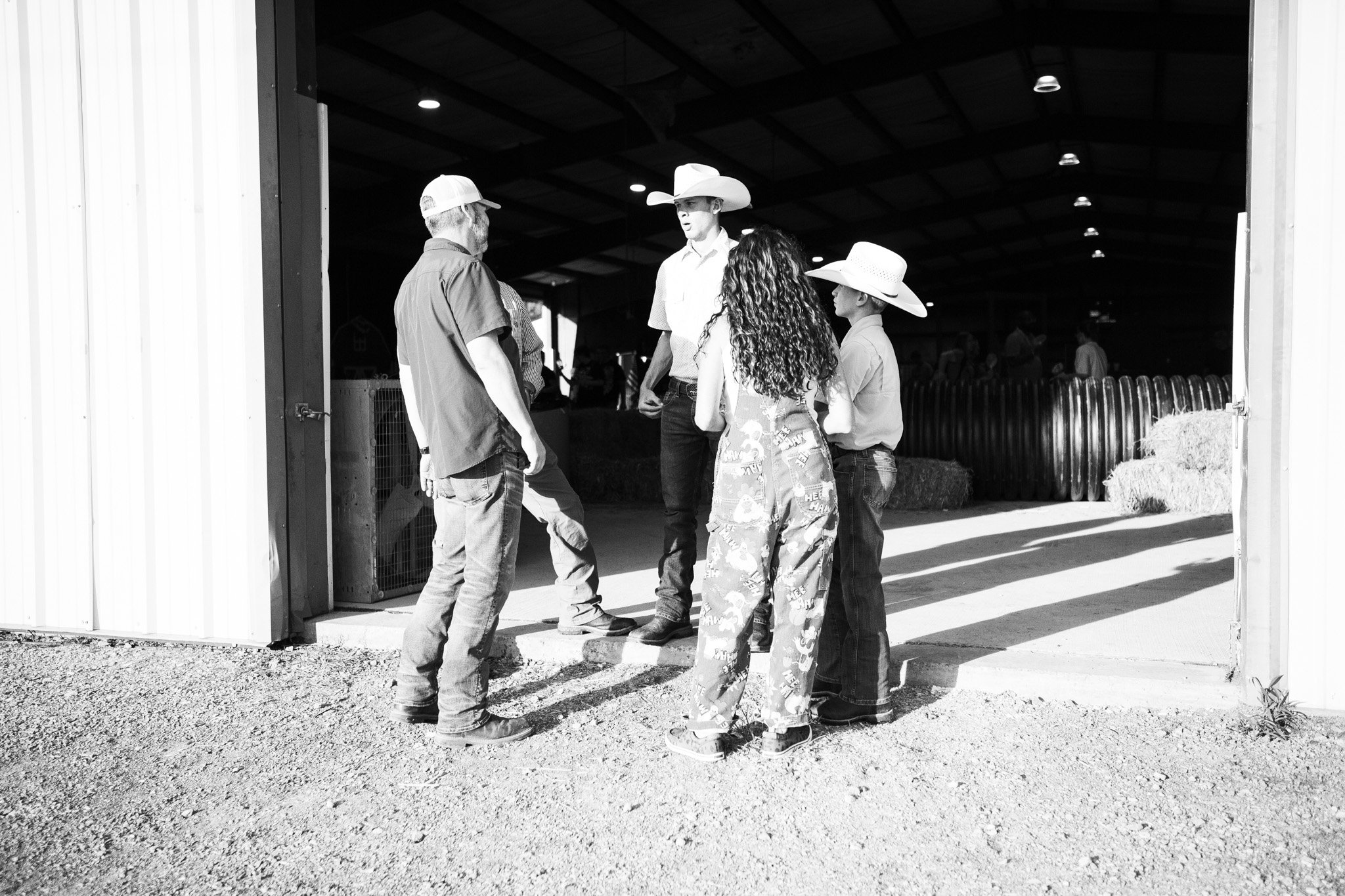 A group of five people, including children and adults, standing at the entrance of a barn, talking. Some are wearing cowboy hats and casual clothing. The barn is open, revealing hay bales inside.