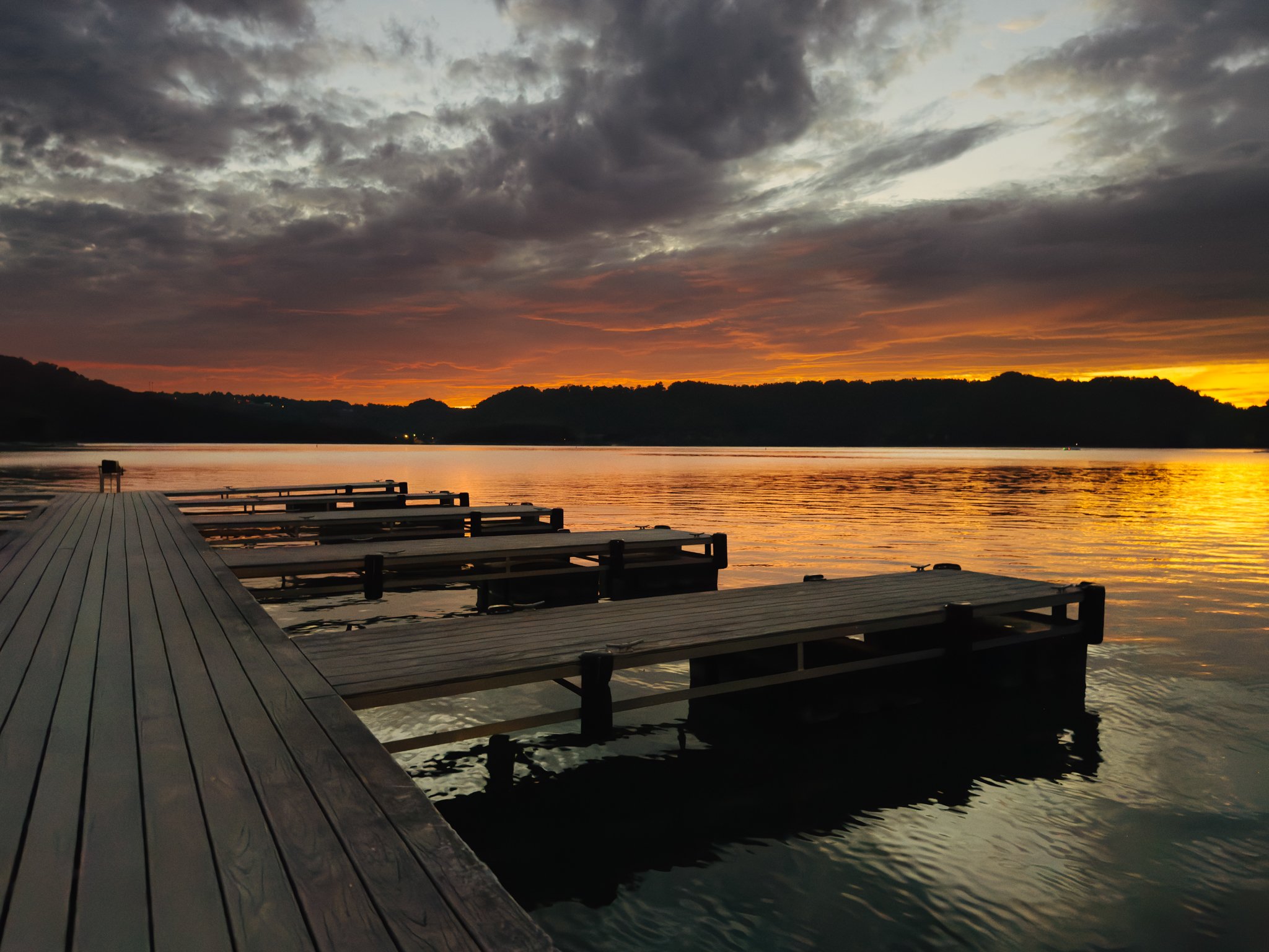 A wooden dock extends over a calm body of water during sunset, with hills in the background and dark, cloudy skies overhead.