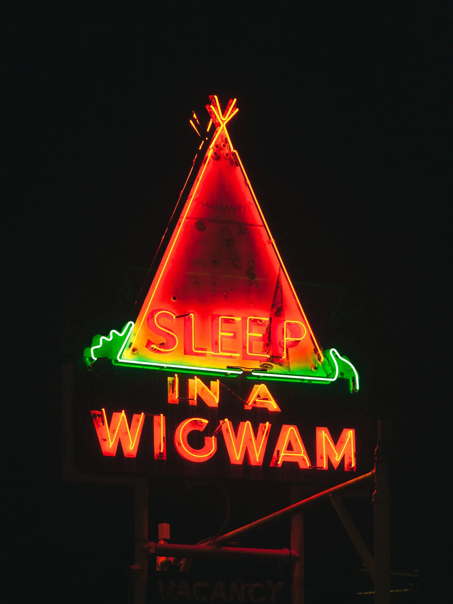 Neon sign with an orange triangle shape, red and yellow text reading 'Sleep in Wigswam', and green accents, against a dark background.