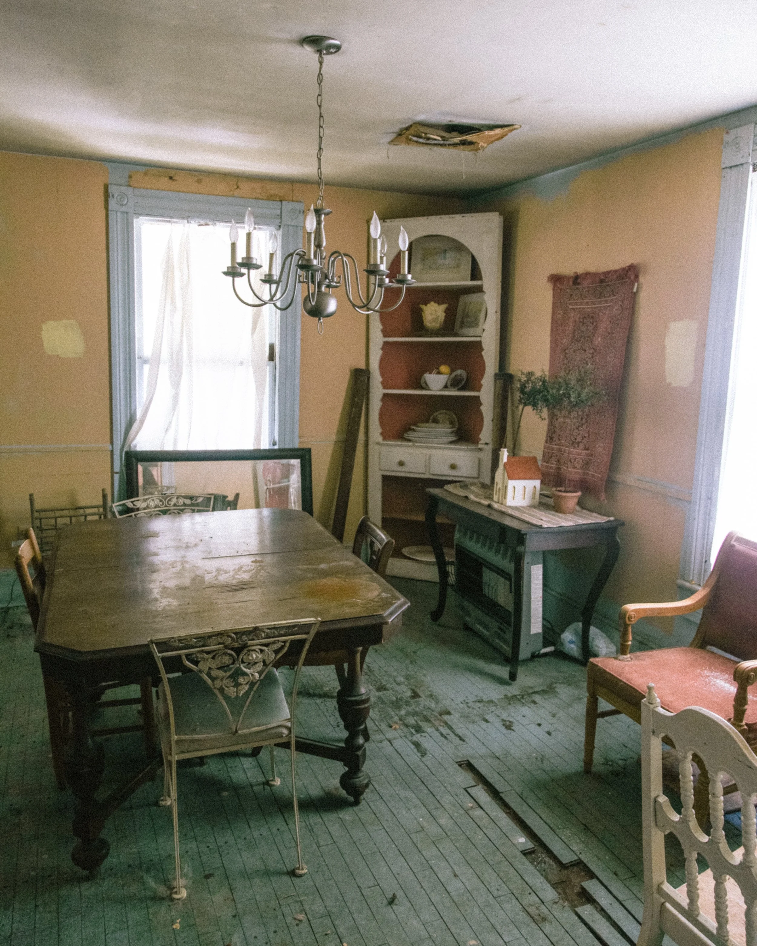 An old dining room with worn and peeling paint, a damaged ceiling with a hole, a large wooden table, mixed chairs including a vintage white one, a chandelier hanging from the ceiling, a white bookshelf with dishes and books, a potted plant, a small s