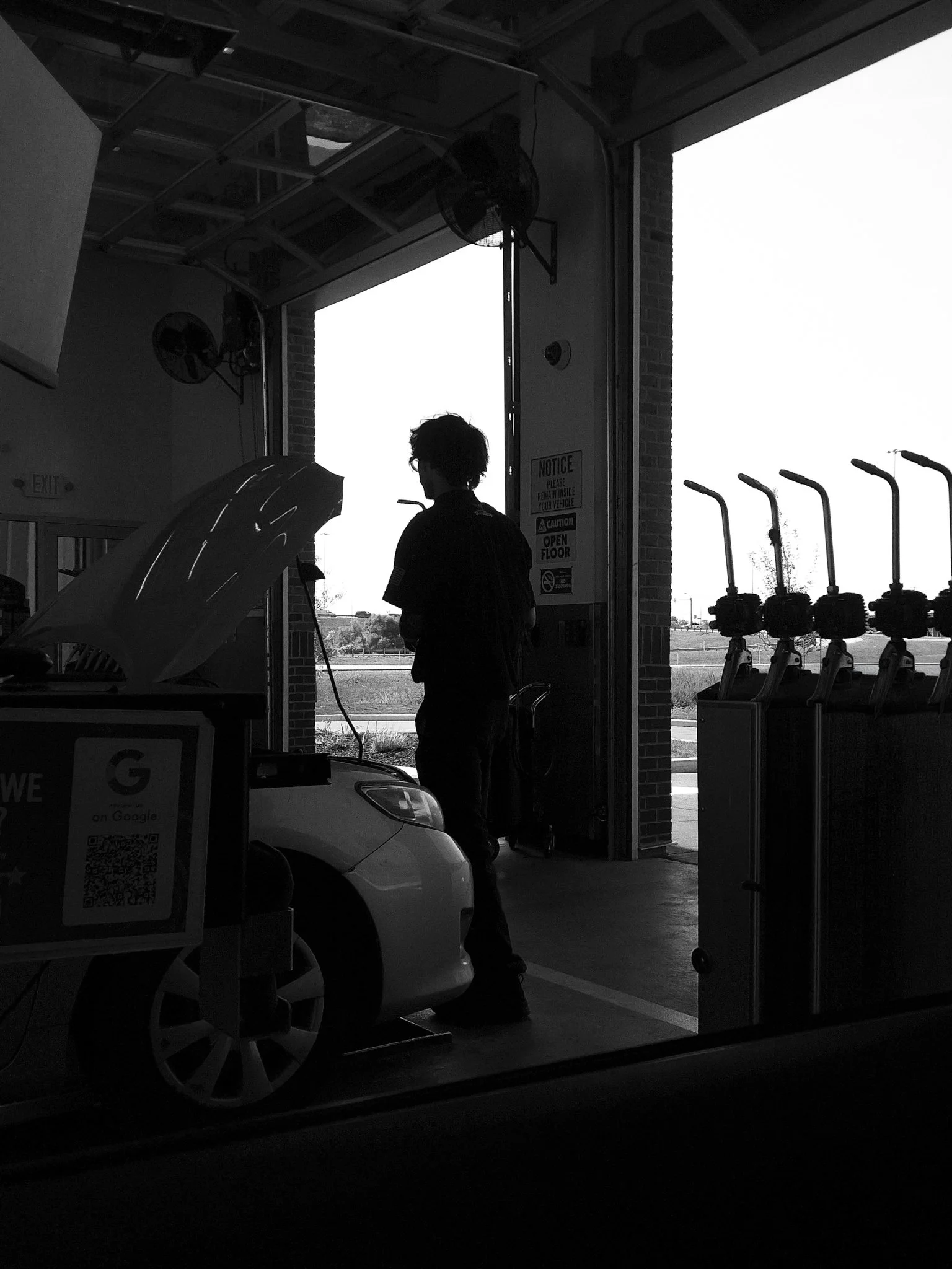A person with curly hair and glasses standing inside a garage or service station, looking outside. There is a white car with an open trunk and several gas pumps visible outside. The image is in black and white.