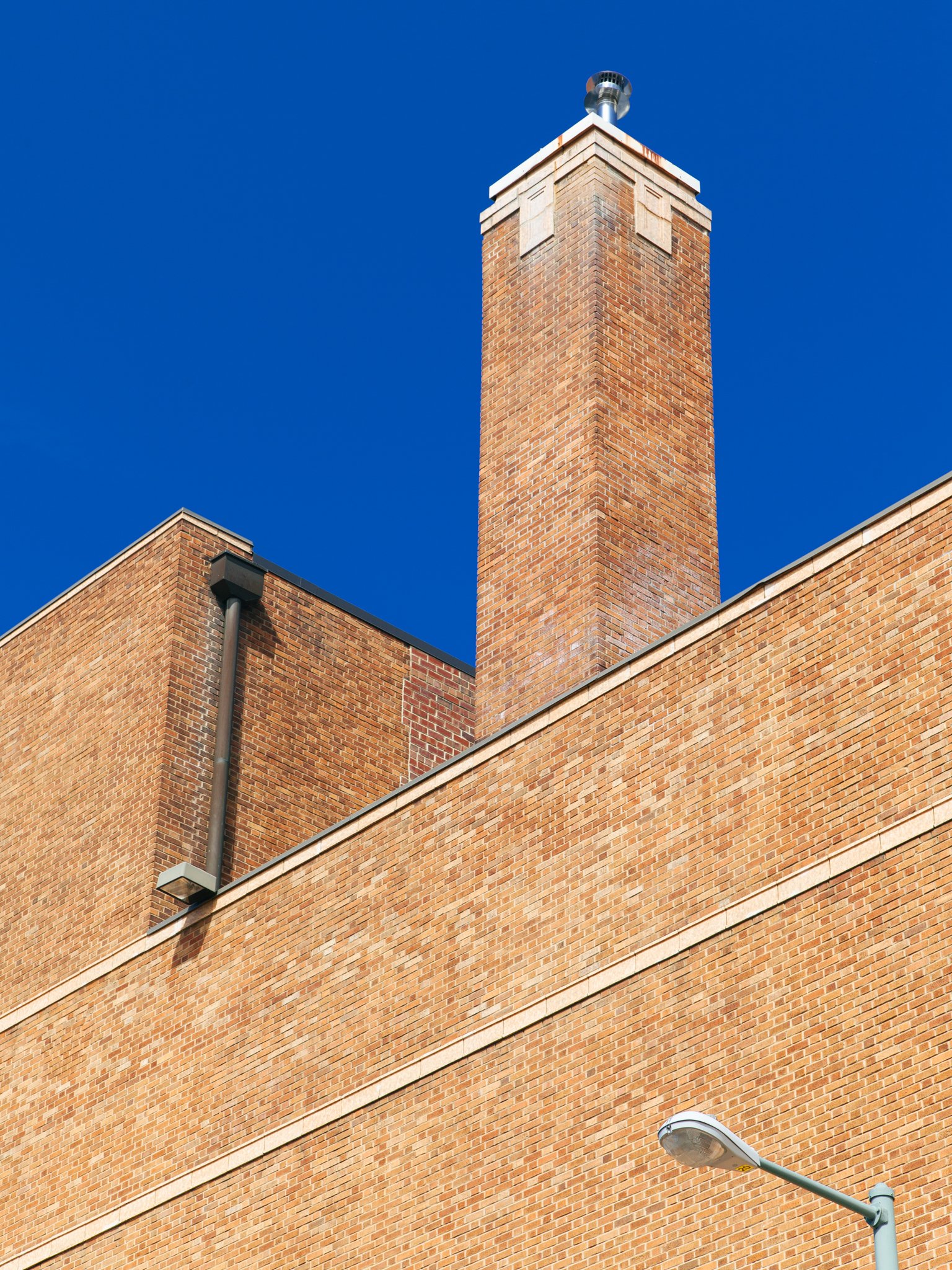 Brick building with a tall brick chimney and a streetlight in the foreground against a blue sky.