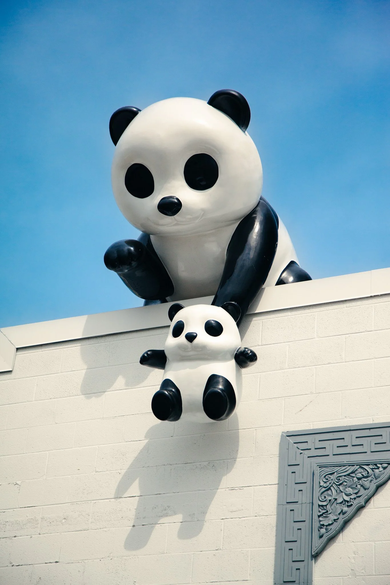 Large panda sculpture sitting on a ledge looking down, with a smaller panda sculpture hanging below on a wall with Chinese decorative art.