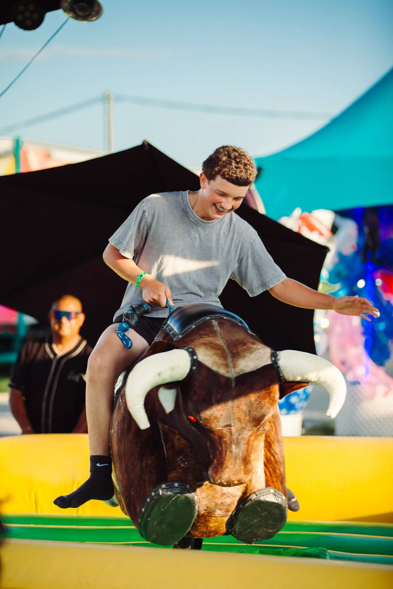 A young person with short curly hair riding a mechanical bull at an outdoor event, smiling widely. In the background, there are colorful tents and a woman wearing sunglasses.