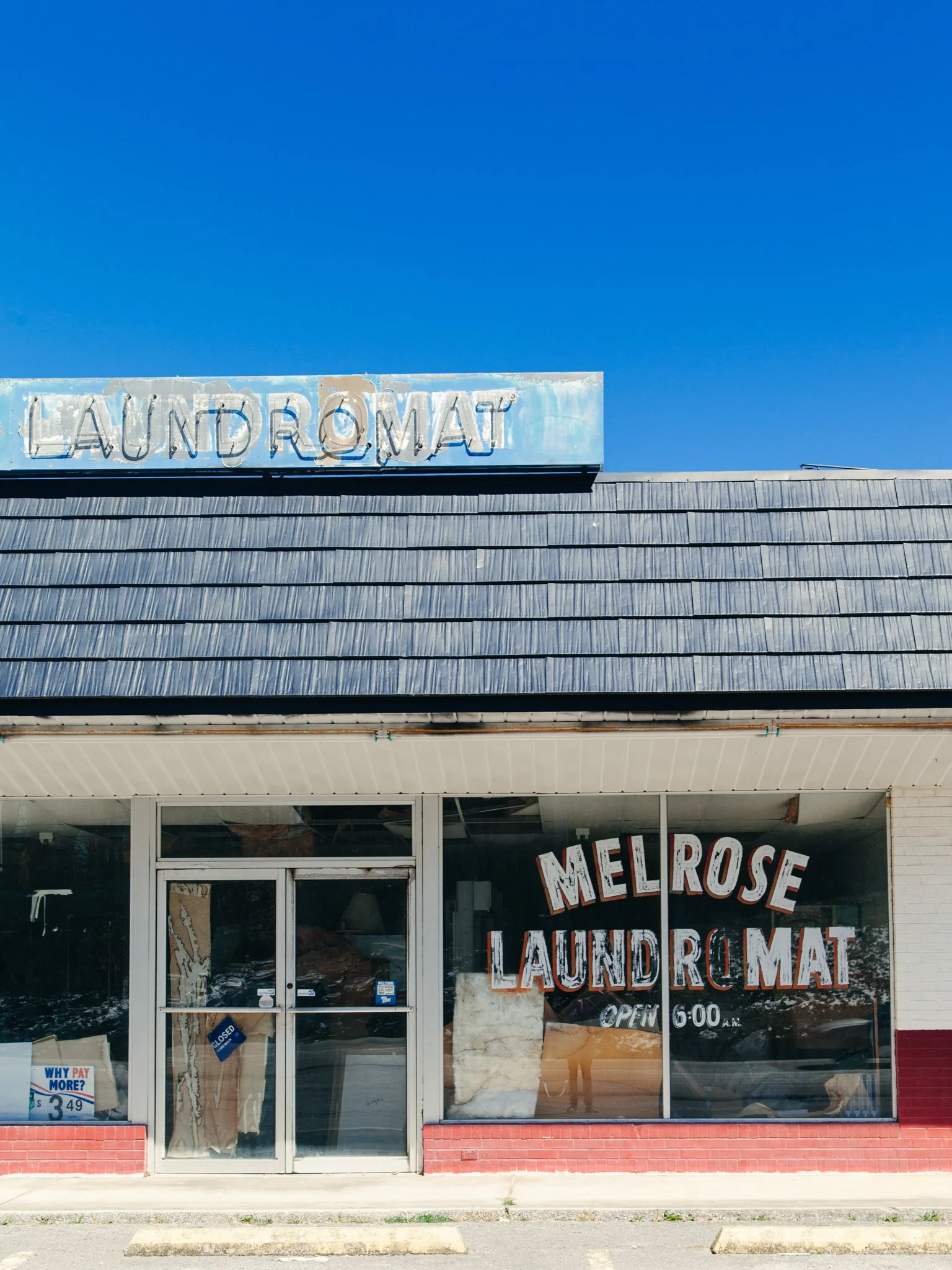 The storefront of a laundromat with a sign that says 'LAUNDROMAT' in neon letters on the roof. The front window displays the words 'MELROSE LAUNDROMAT' and an open sign. The building has a corrugated metal awning and glass doors.