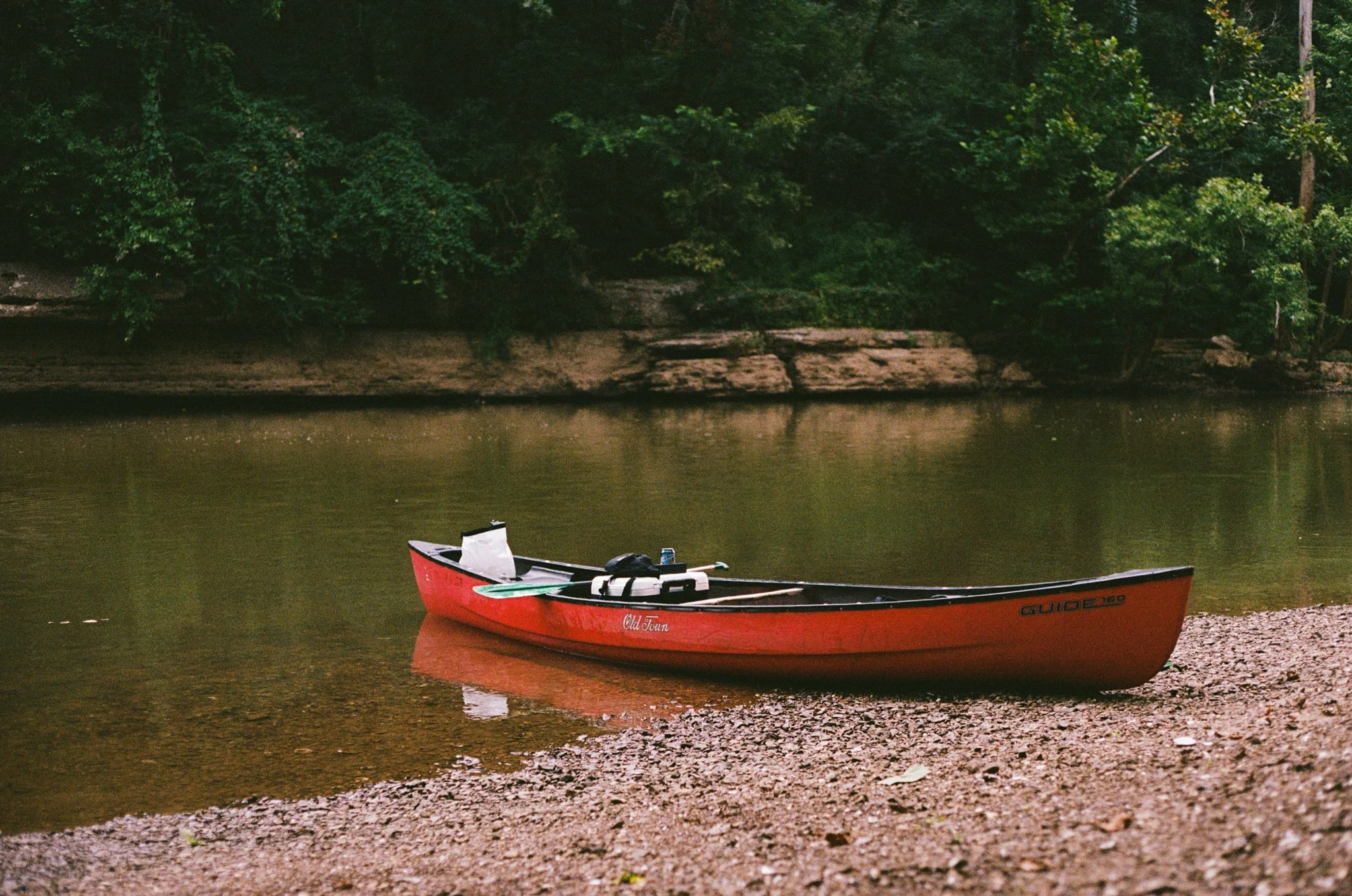 A red canoe with a white interior resting on a rocky shore next to a calm body of water, surrounded by lush green trees.