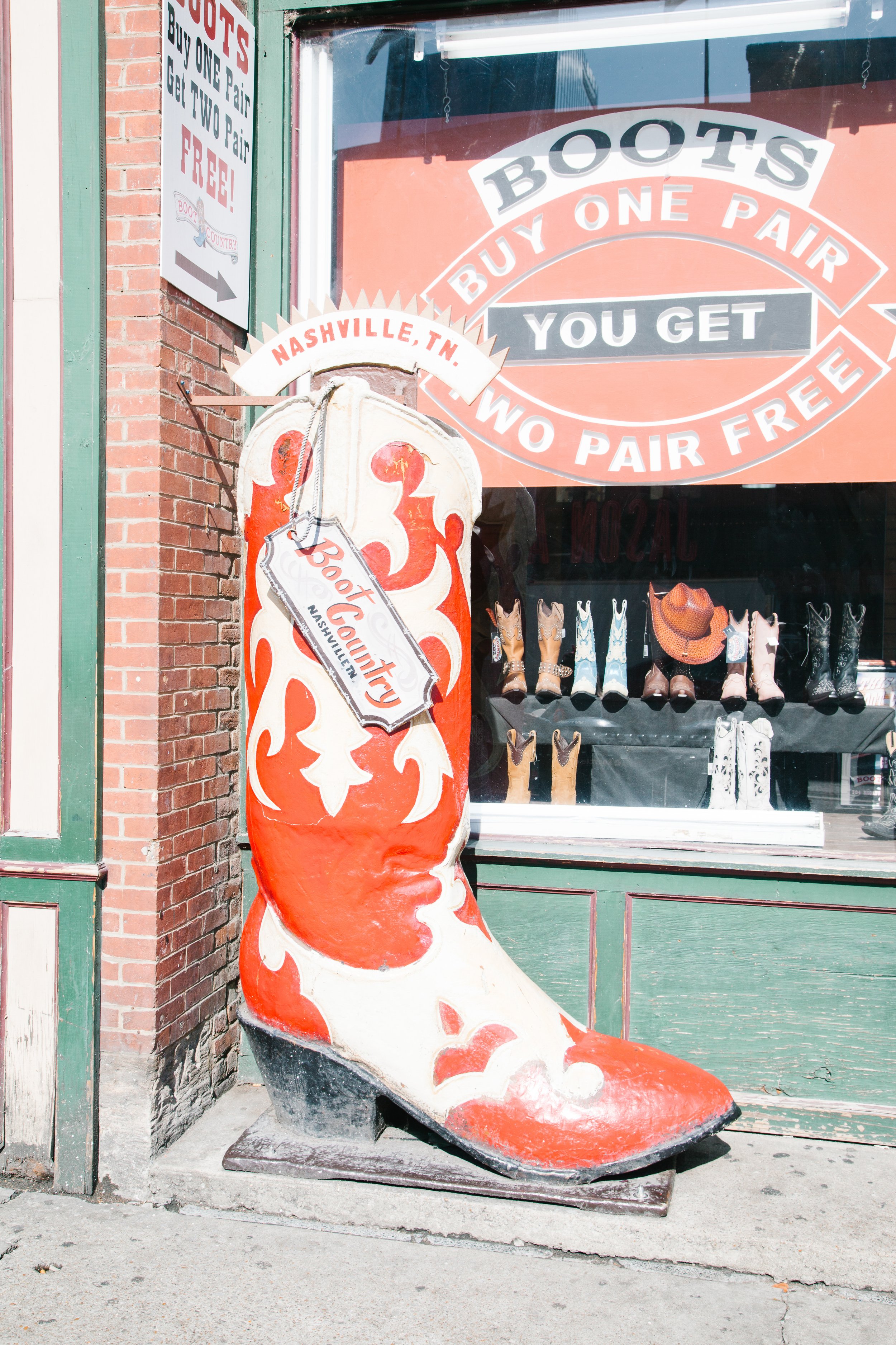 A large cowboy boot sculpture painted to resemble a red, orange, and white flame pattern, placed outside a store. The storefront window displays cowboy boots and hats, with signs advertising buy one, get one free offers and indicating the store is in