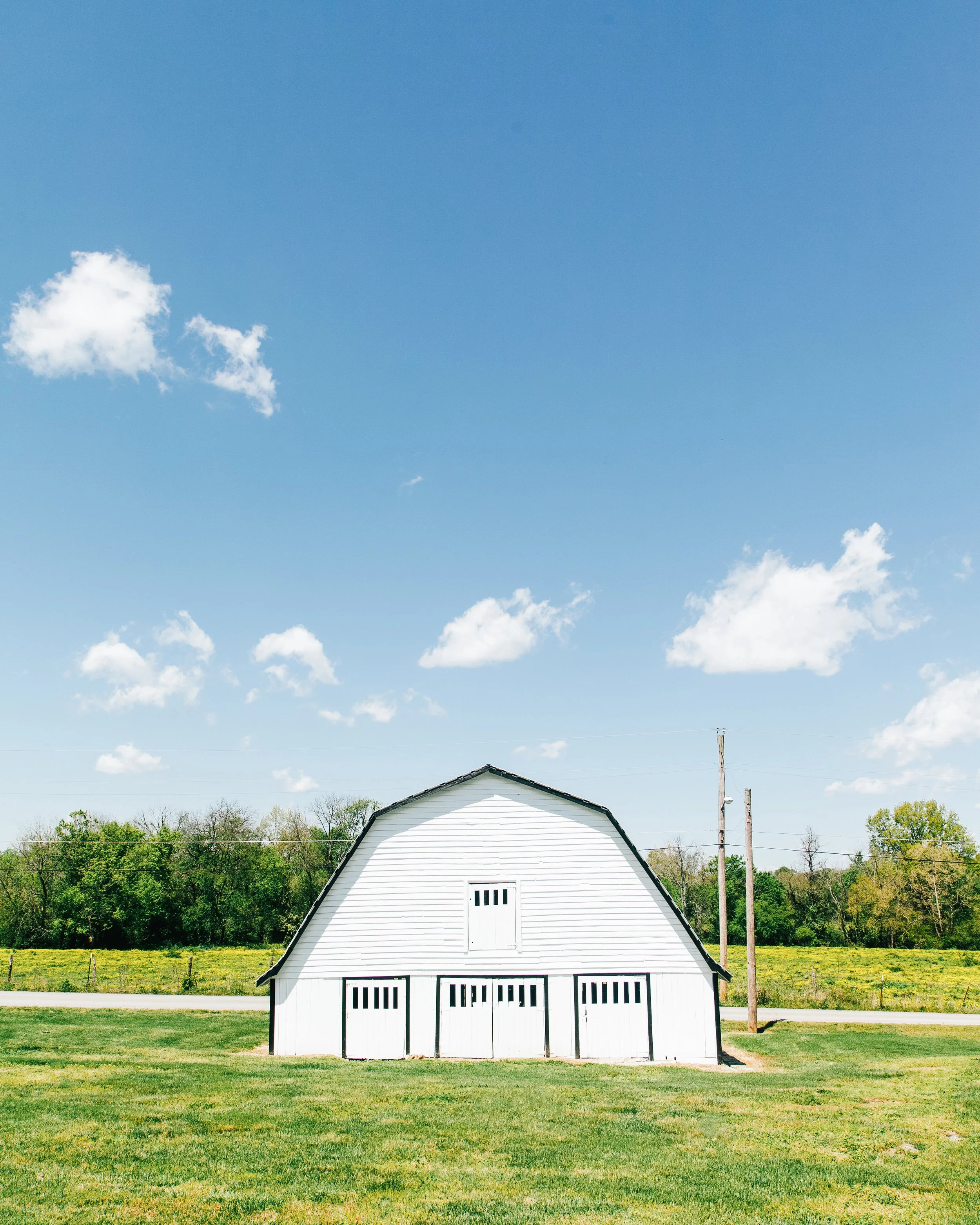 A white barn with black trim on a grassy field, with trees, utility poles, and a blue sky with scattered clouds in the background.