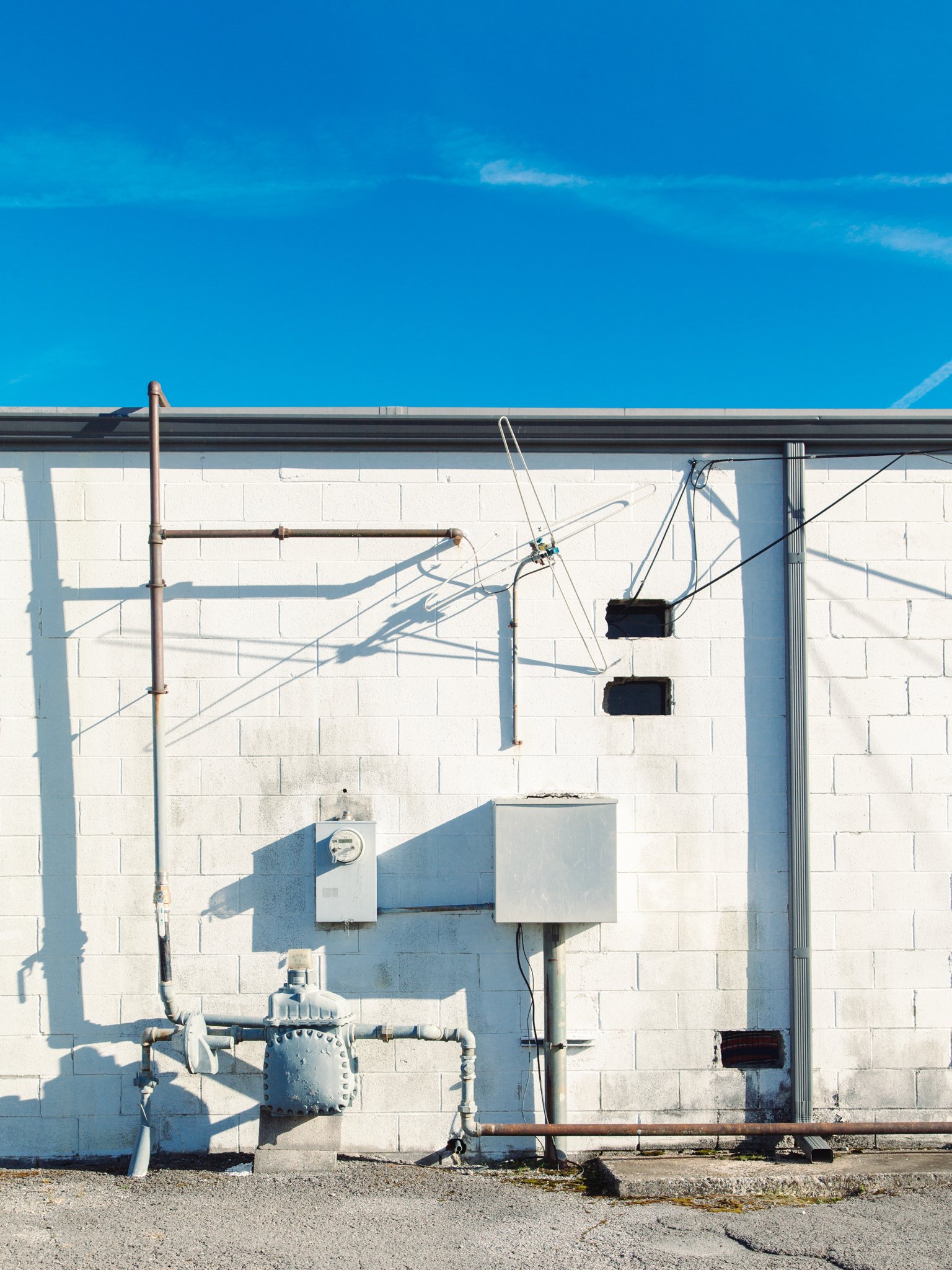 Exterior view of a white brick building's wall with visible pipes, electrical meters, and vents against a bright blue sky.