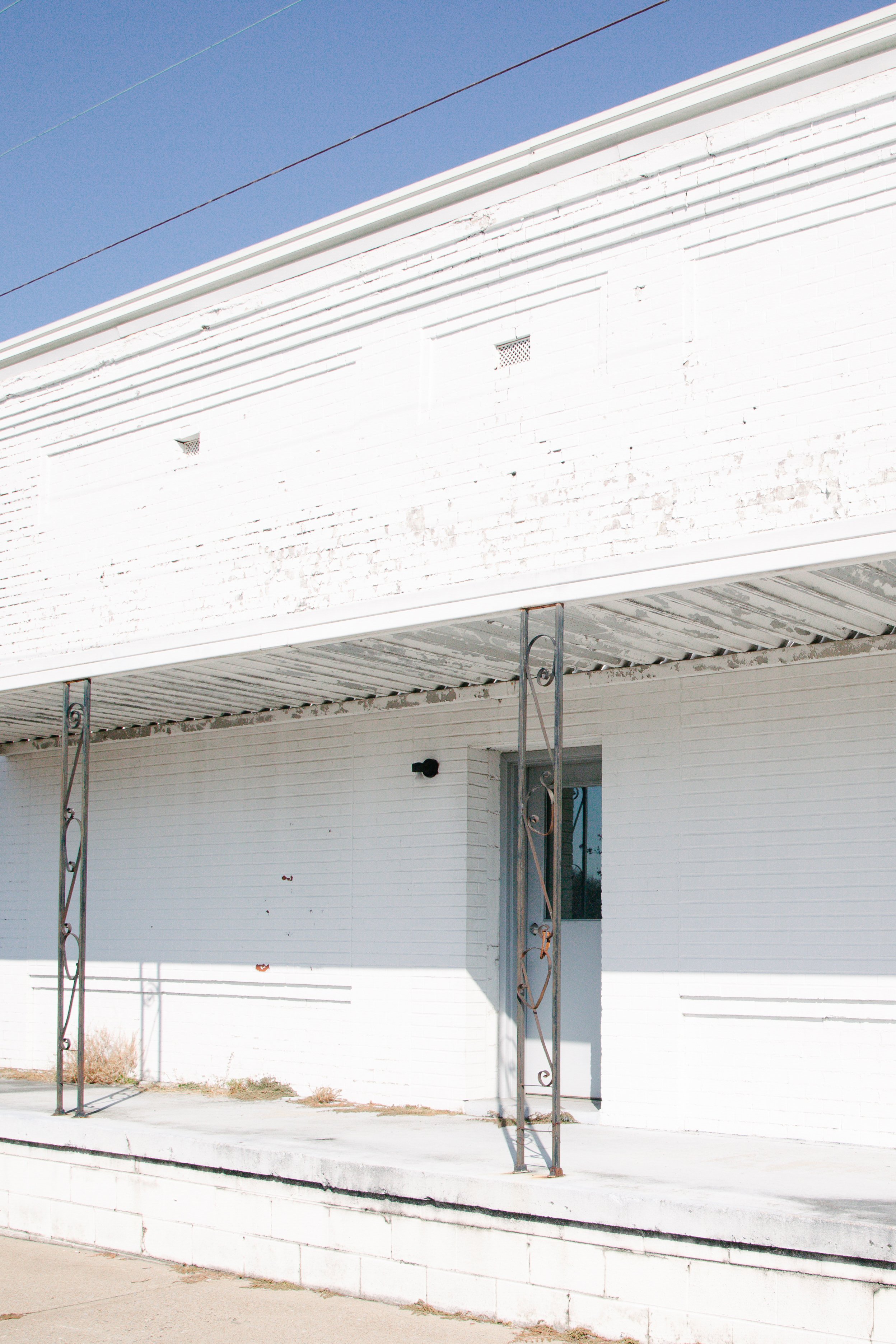 A white building with a balcony and iron support columns, two small square vents, and a door. The wall has some discoloration and damage. The balcony has a white railing and a corrugated roof, with wires above. The sky is blue.