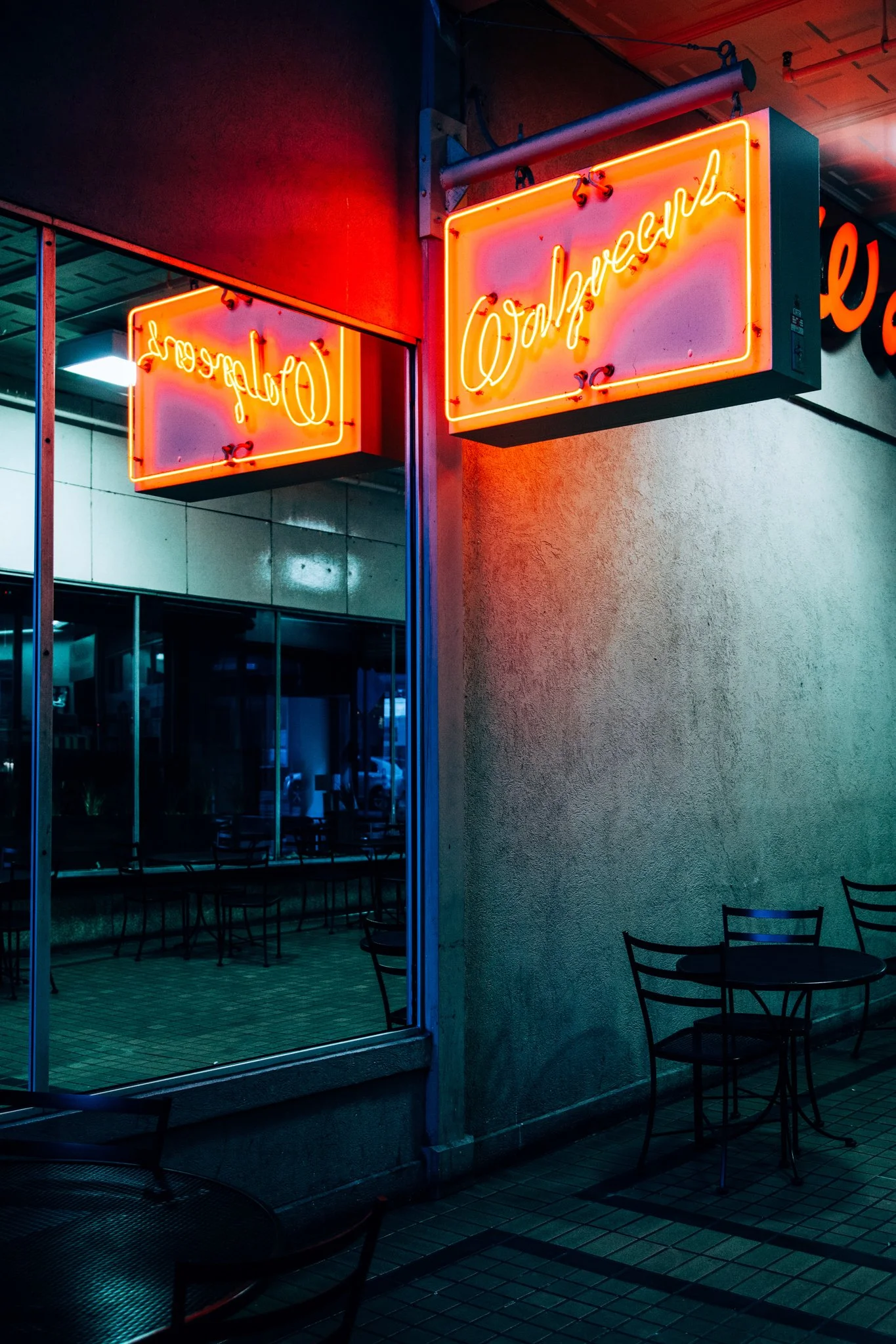 Close-up of illuminated neon sign with cursive text in orange, reflected in a glass window at night, outside a building with a textured wall and seating area.