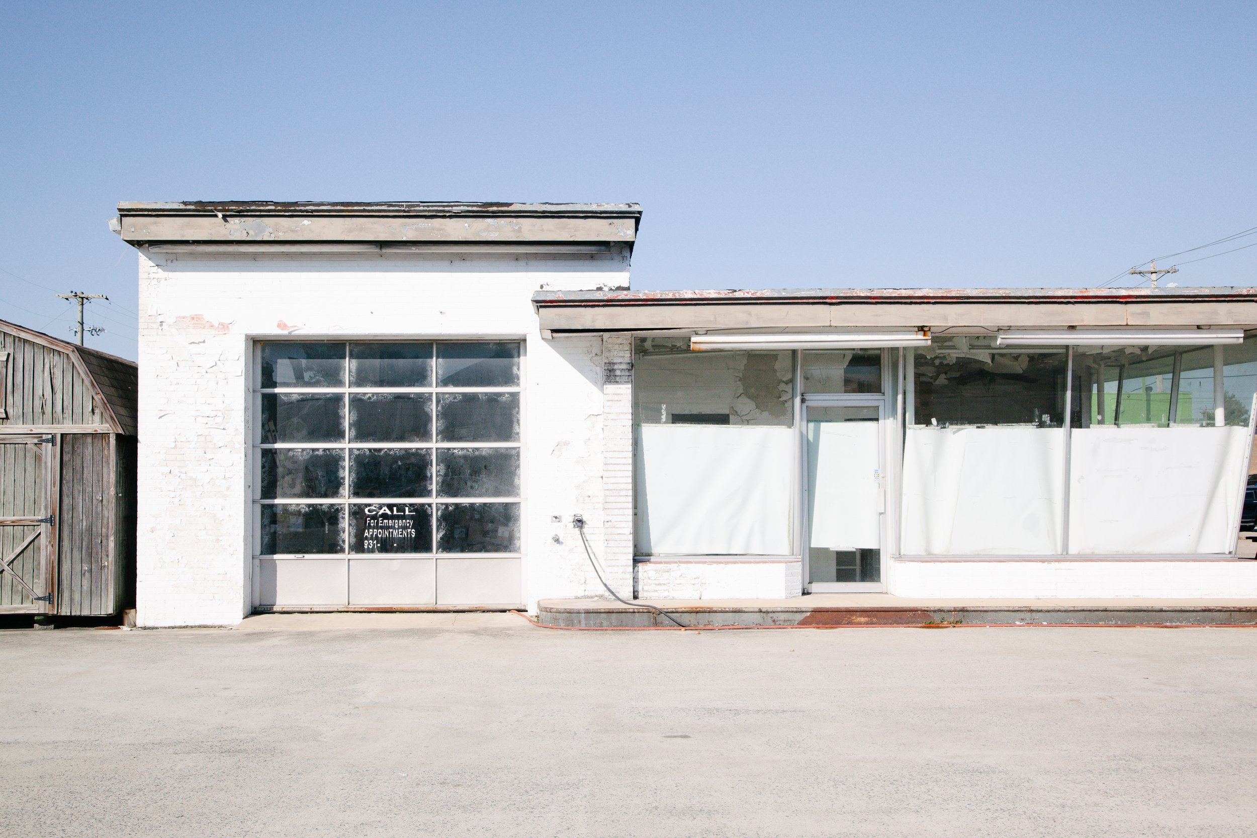 An old, white brick building with a large glass window and a smaller glass entry door. The window has a sign that reads 'CALL for emergency appointments.' The building appears to be in disrepair with peeling paint and a damaged roof. There is a woode