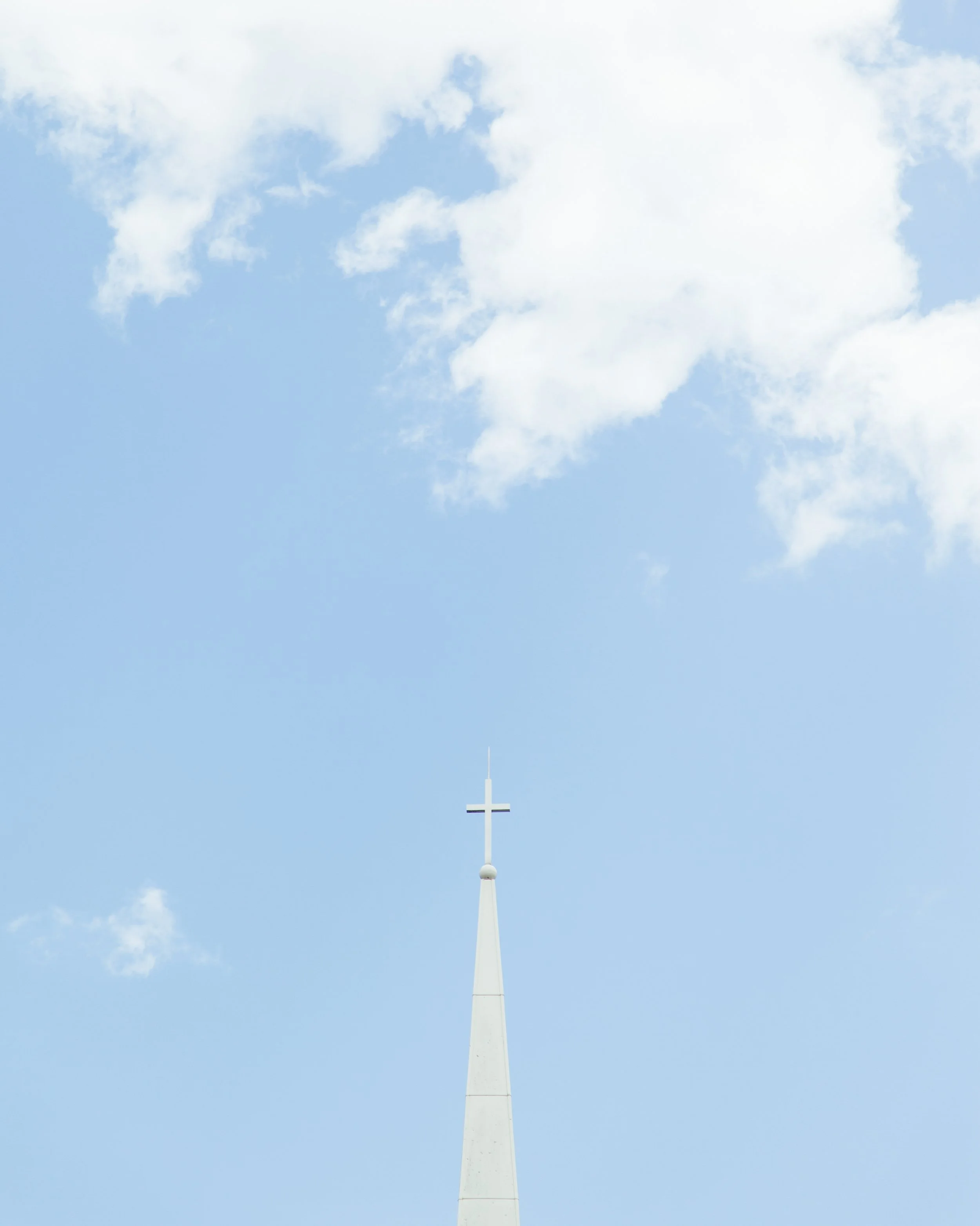A white cross atop a tall white steeple reaching into a blue sky with scattered white clouds.