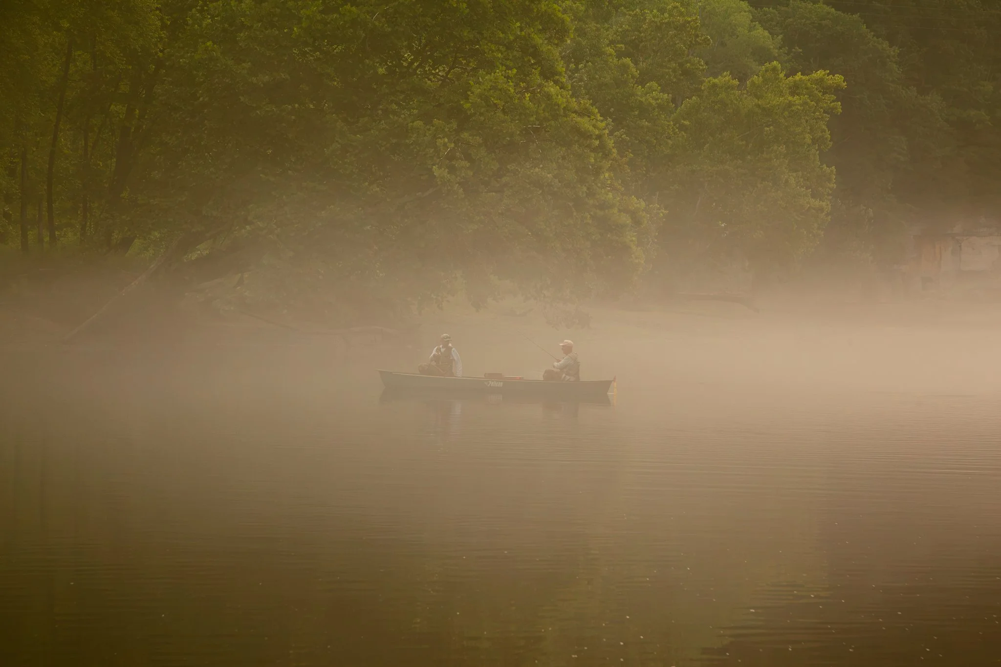 Two people fishing from a boat on a calm river surrounded by dense green trees, with mist or fog on the water.