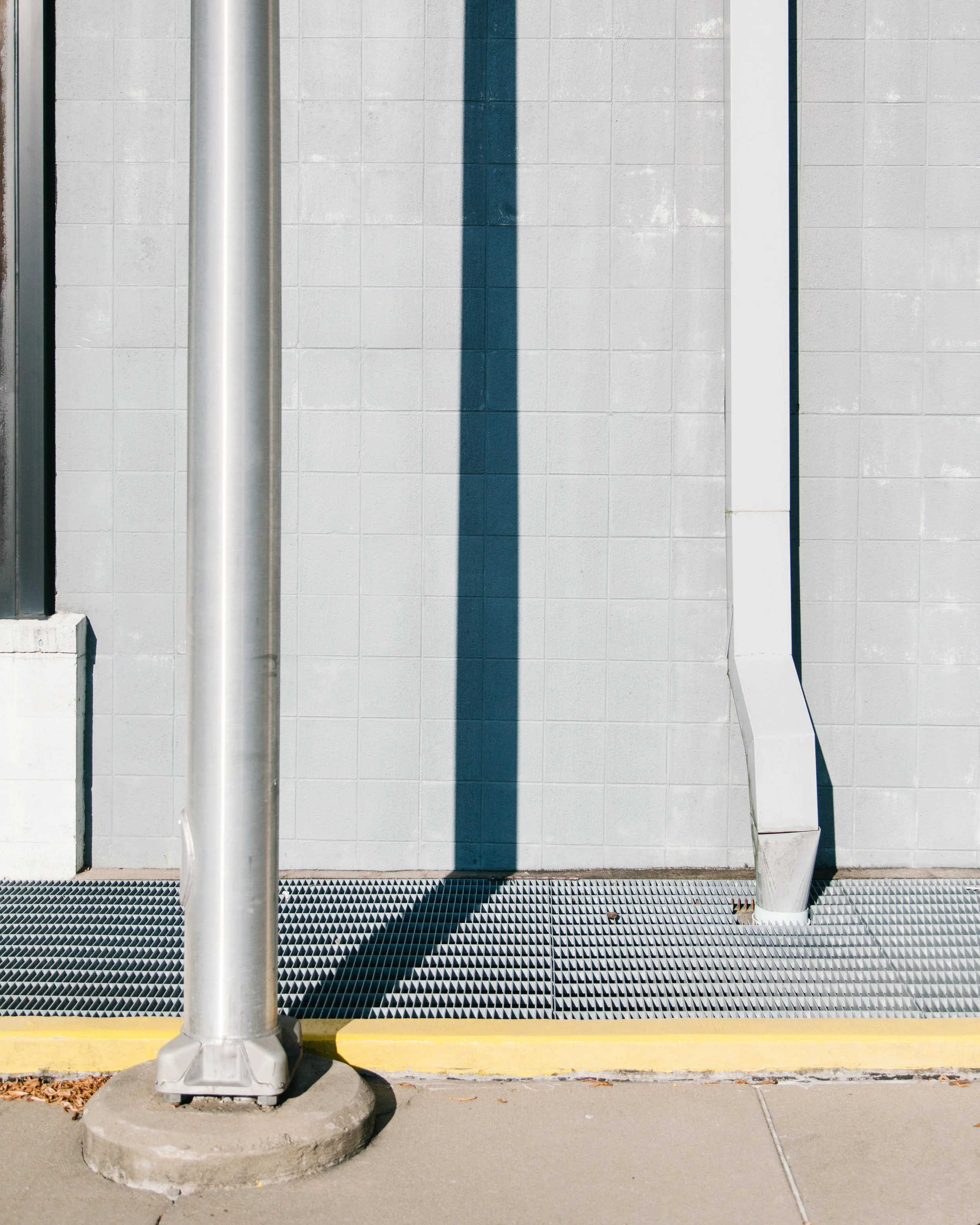 Photo of a metal pole casting a shadow on a tiled wall, with a grate on the ground and a white pipe on the right side.