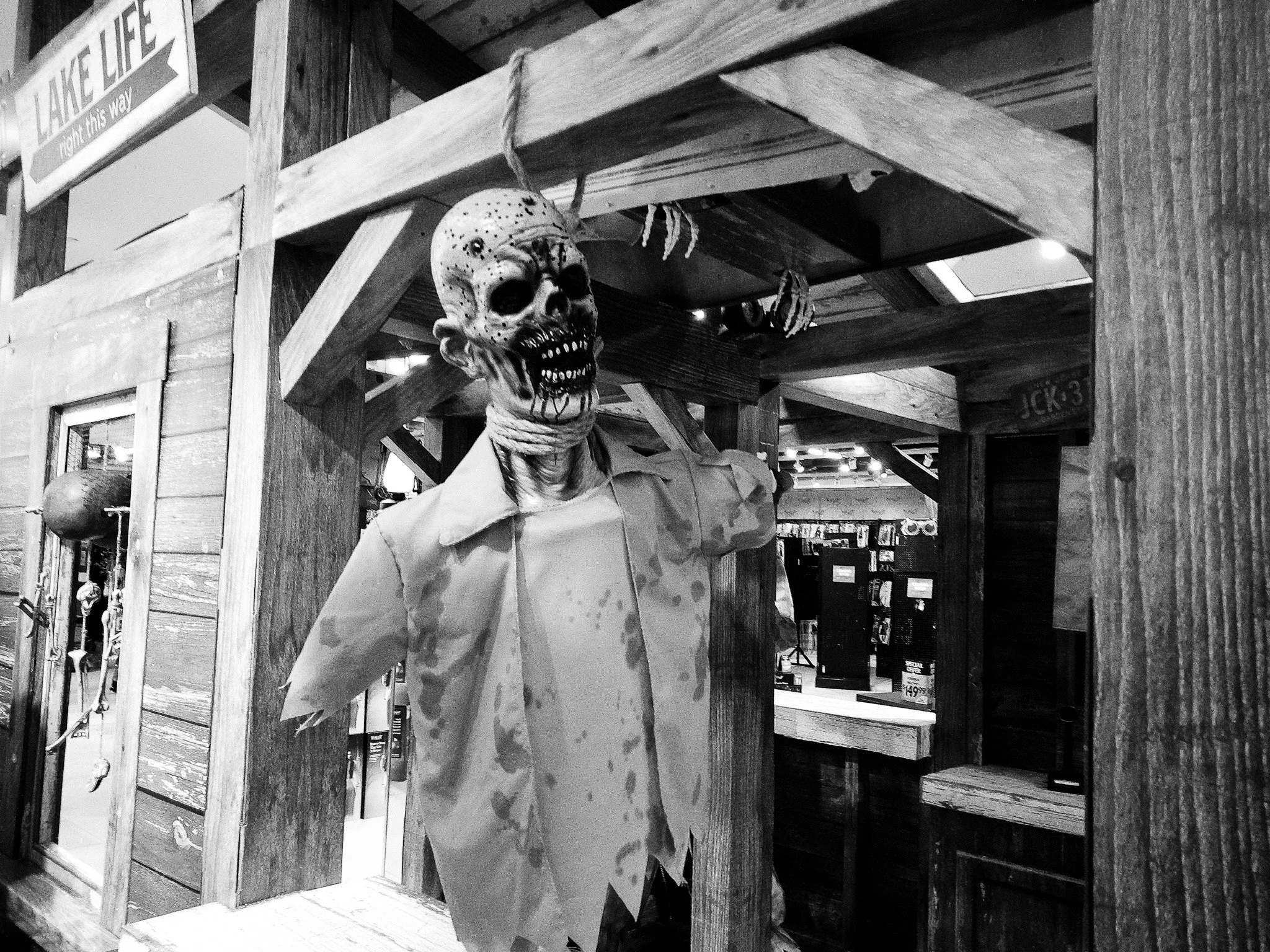 A Halloween-themed display featuring a skeleton wearing a mask that looks like a hockey mask, a white shirt, and a tie. The skeleton is positioned behind a rustic wooden structure with a sign that reads 'LAKE LIFE' and 'right this way.' The backgroun